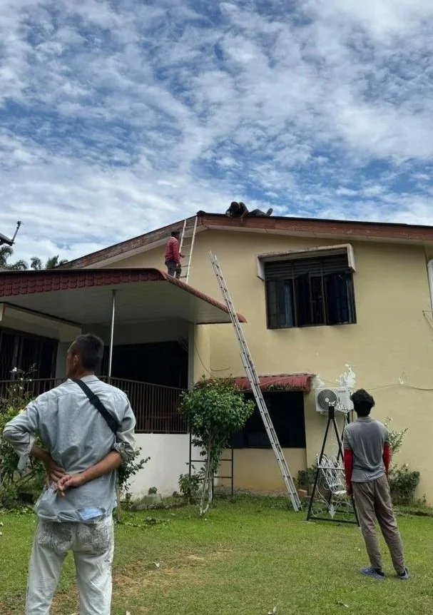 Three men near a yellow house, with one on the roof, one on the ladder, and one standing on the ground watching, under a partly cloudy sky.