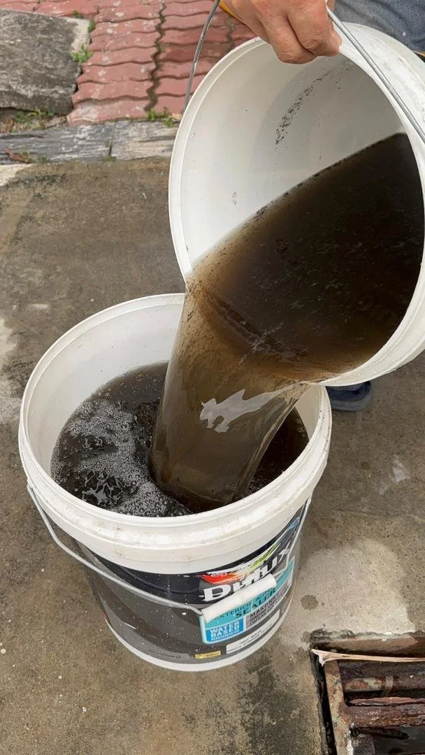 A person pouring a black liquid, possibly motor oil or dirty water, from a bucket into another container outdoors on a concrete surface.