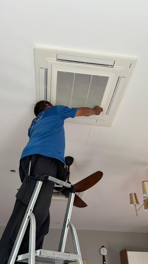 A technician standing on a ladder adjusting a ceiling air conditioning vent in a room.