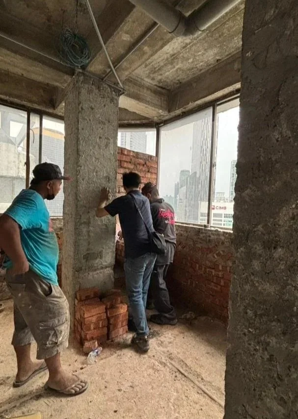 Three men standing inside an unfinished building with brick and concrete walls, looking out the large window at the cityscape outside, with construction materials on the floor.
