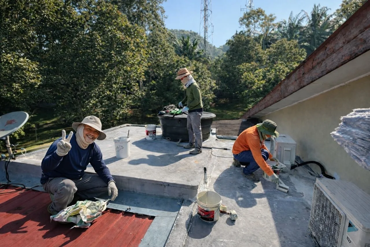 Three workers on a rooftop doing maintenance or construction work, with one smiling and giving a peace sign, and others installing or repairing an air conditioning unit and handling tools, surrounded by trees and clear sky.