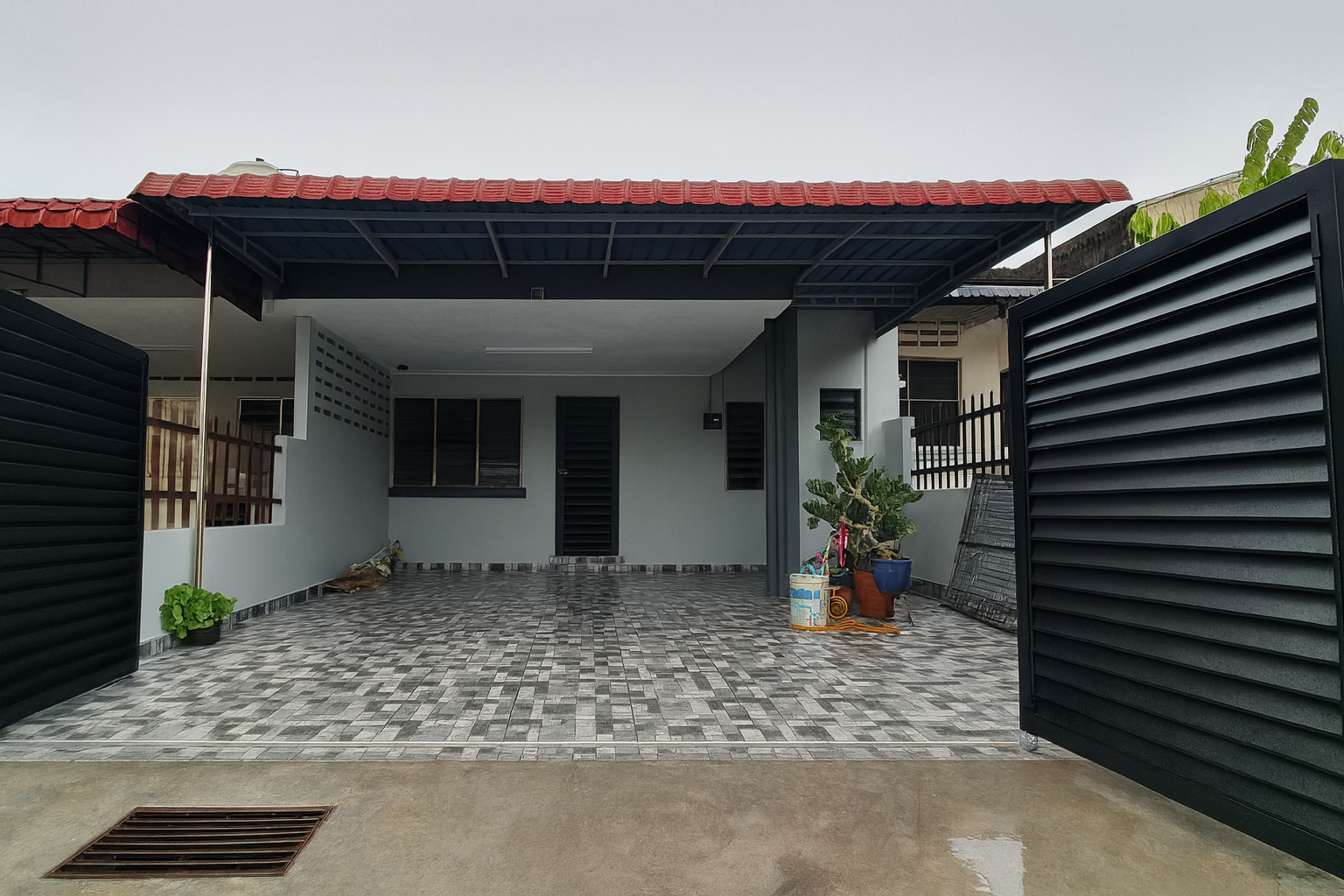 View of a modern garage with black sliding gates, a tiled driveway, and potted plants near the entrance, located in front of a house with white walls and a red tiled roof.