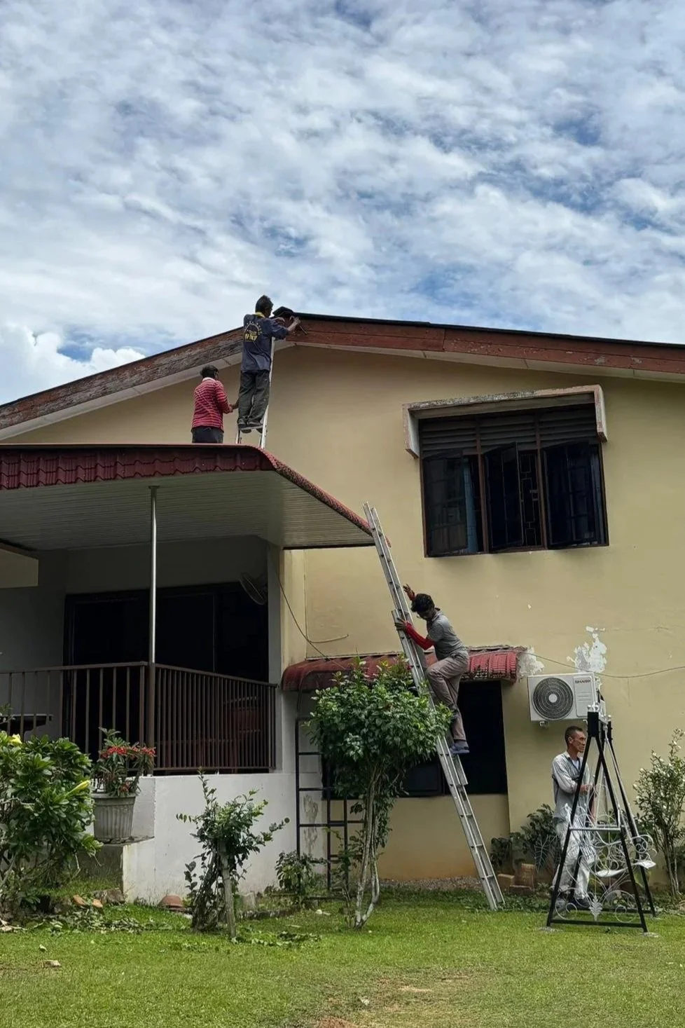 People working on the roof of a house with ladders, one person is holding a tool and another is standing nearby observing, while a fourth person stands on the ground with a tripod.