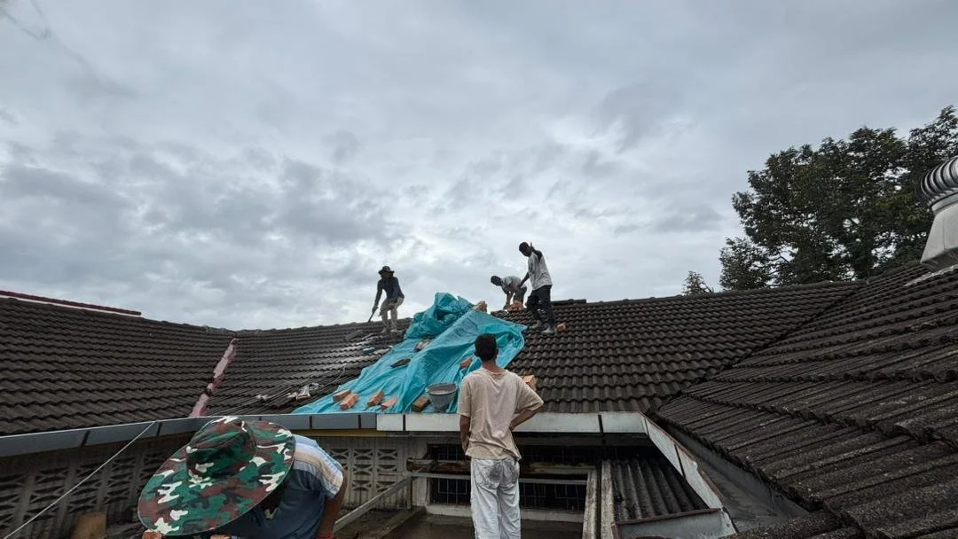 Four workers on a roof, with some damage or construction work happening, covered with a blue tarp, under a cloudy sky.