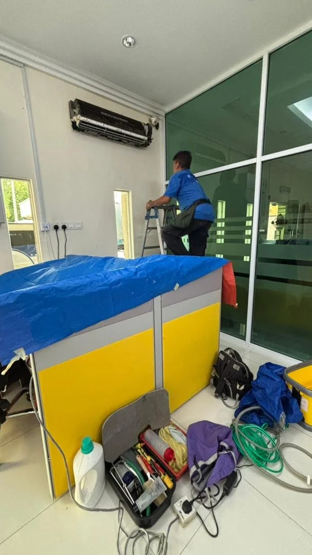 Technician on ladder servicing wall-mounted air conditioning unit in office with cleaning supplies and equipment on the floor.