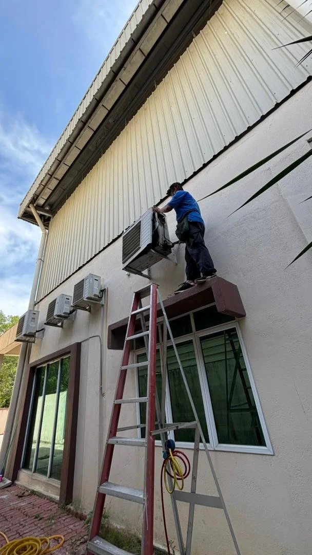 A person standing on a ladder repairing or maintaining an air conditioning unit mounted on the exterior wall of a building.
