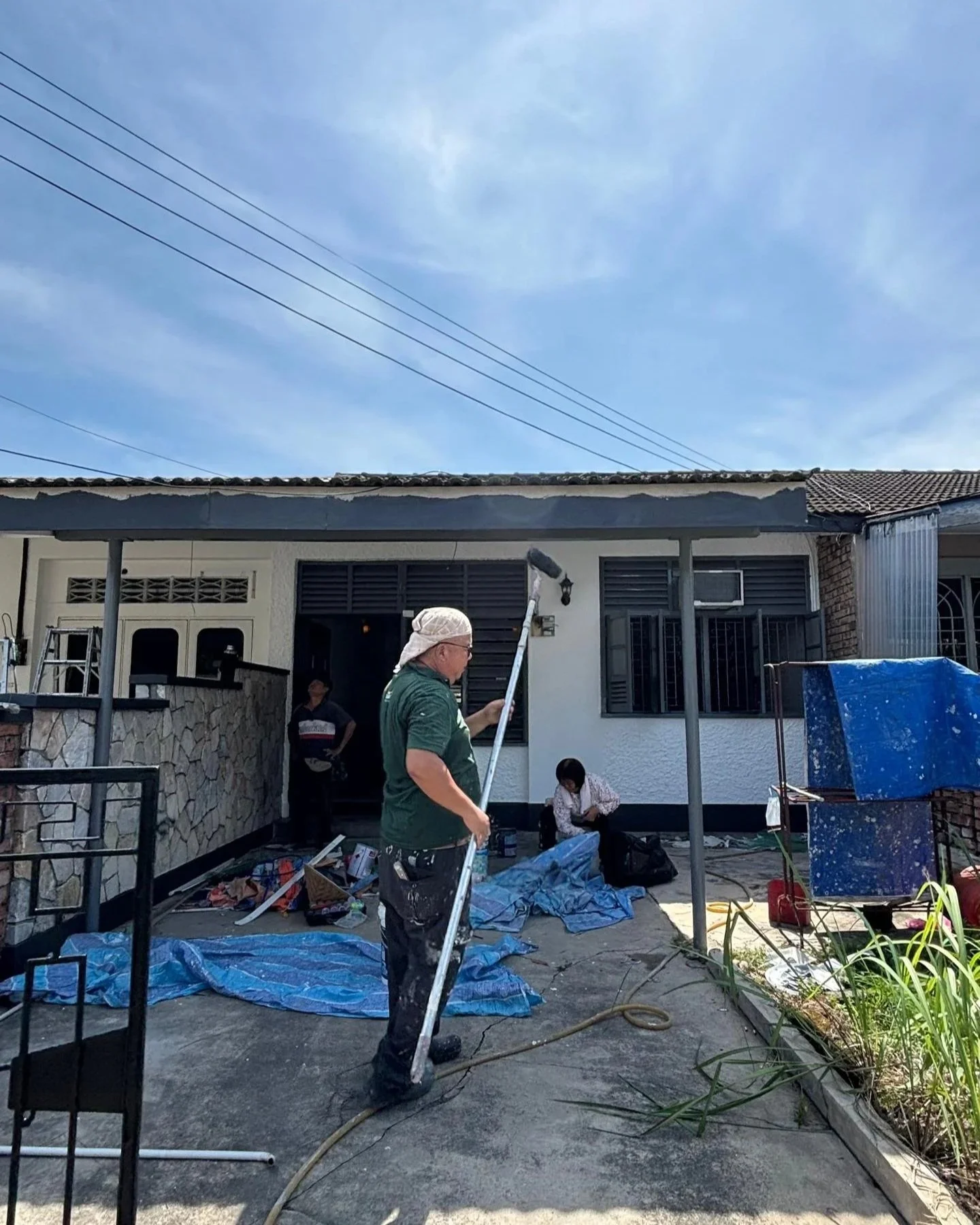 Workers painting the exterior of a house on a sunny day, with one person using a roller brush and others organizing supplies.