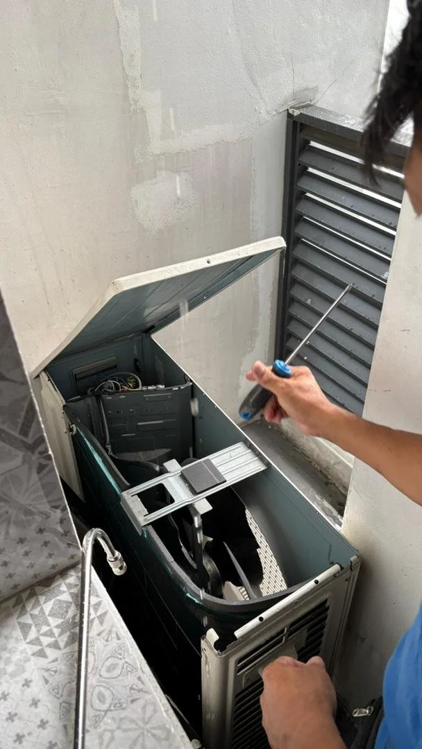 Person repairing or inspecting an open window air conditioning unit with a screwdriver, indoors near a wall with a vent.