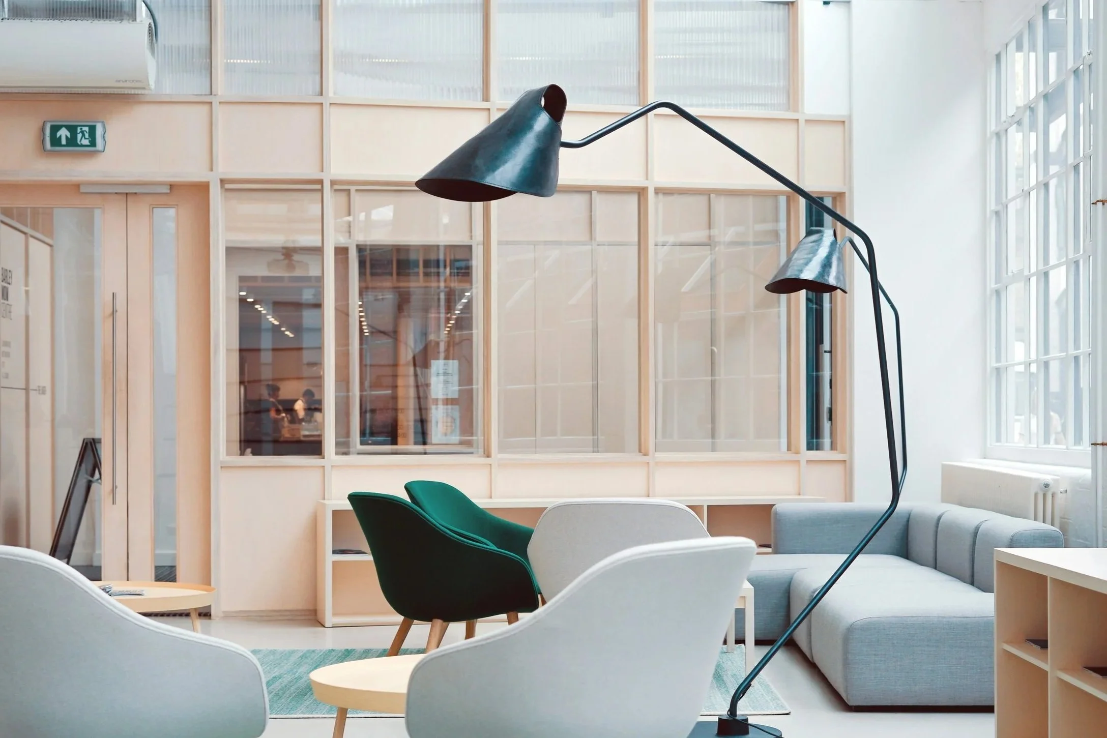 Modern lounge with white and green chairs, a large gray sofa, a black floor lamp, and large windows letting in natural light.