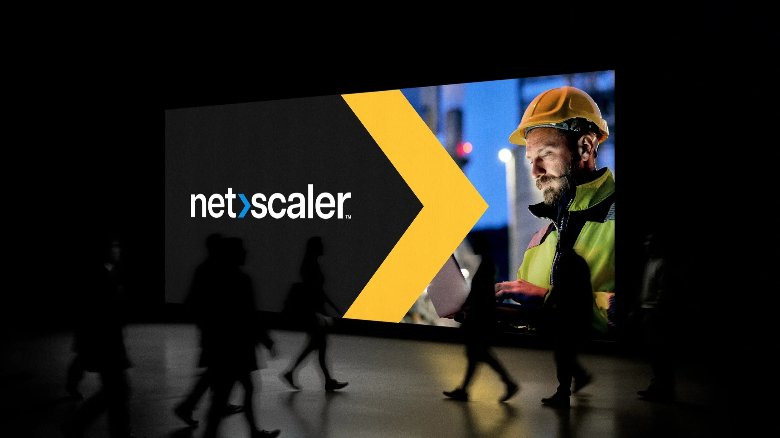 Large illuminated billboard displays the Netscaler logo with a photo of a construction worker wearing a yellow hard hat, a yellow high-visibility vest, and working on a laptop, with several walking silhouettes in front of it.