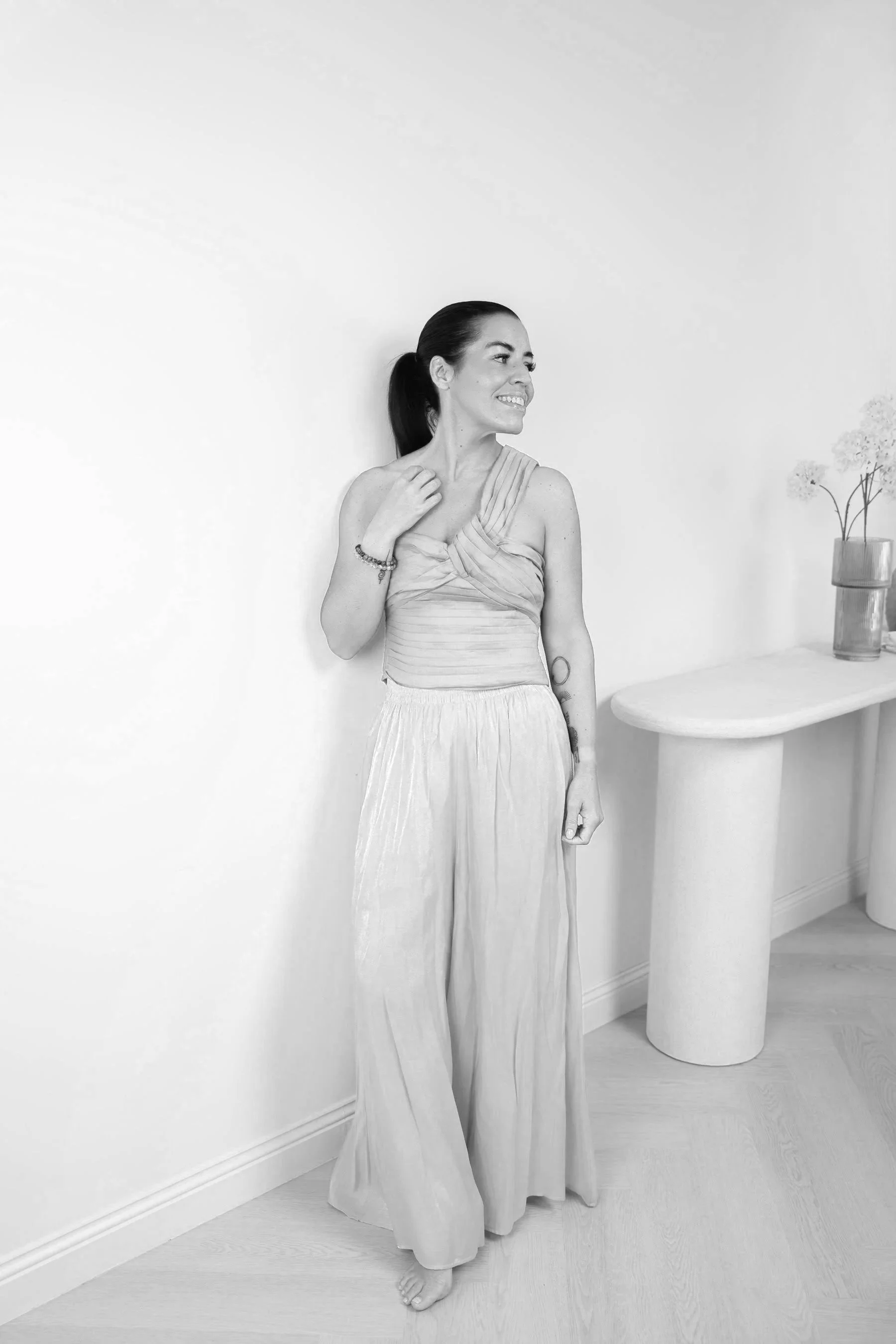 Hayley Mclaren founder of Inner Architect Wellness and Health Coaching, standing up against a white wall wearing light trousers and a one shoulder top with a table in the background