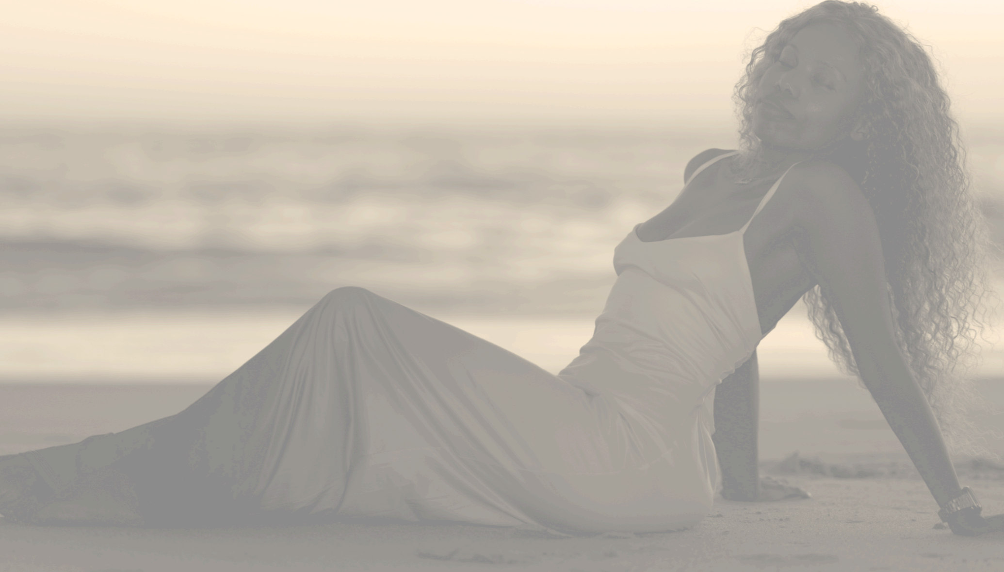 Woman relaxing on the beach during sunset, sitting on sand with the ocean in the background.