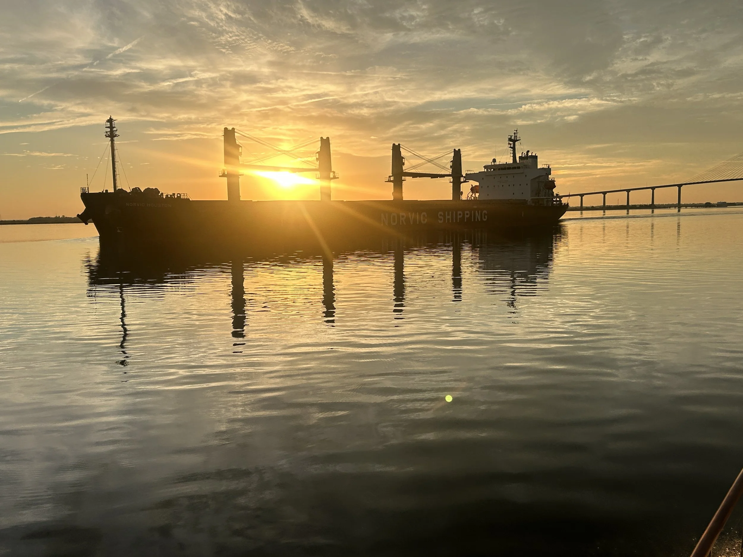 A ship labeled 'Norvic Shipping' on calm water at sunset with the sun setting behind the ship, creating a silhouette effect, along with a distant bridge visible on the right side.