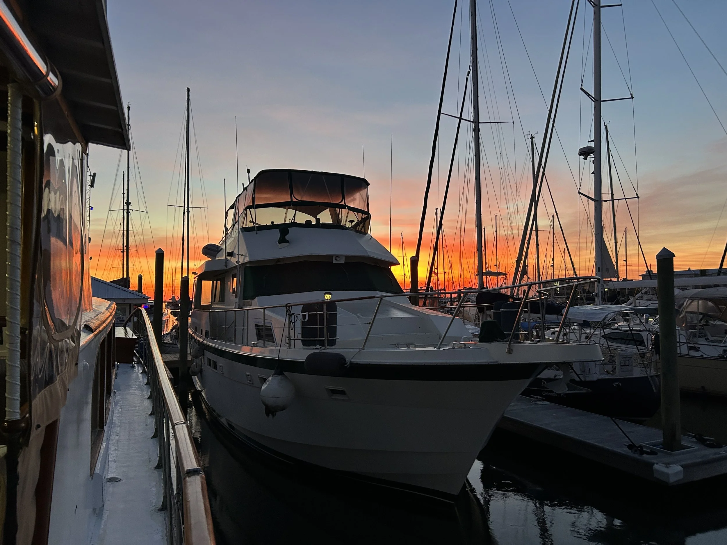 A marina during sunset with multiple sailboats and a yacht docked at the pier, casting reflections on the water.