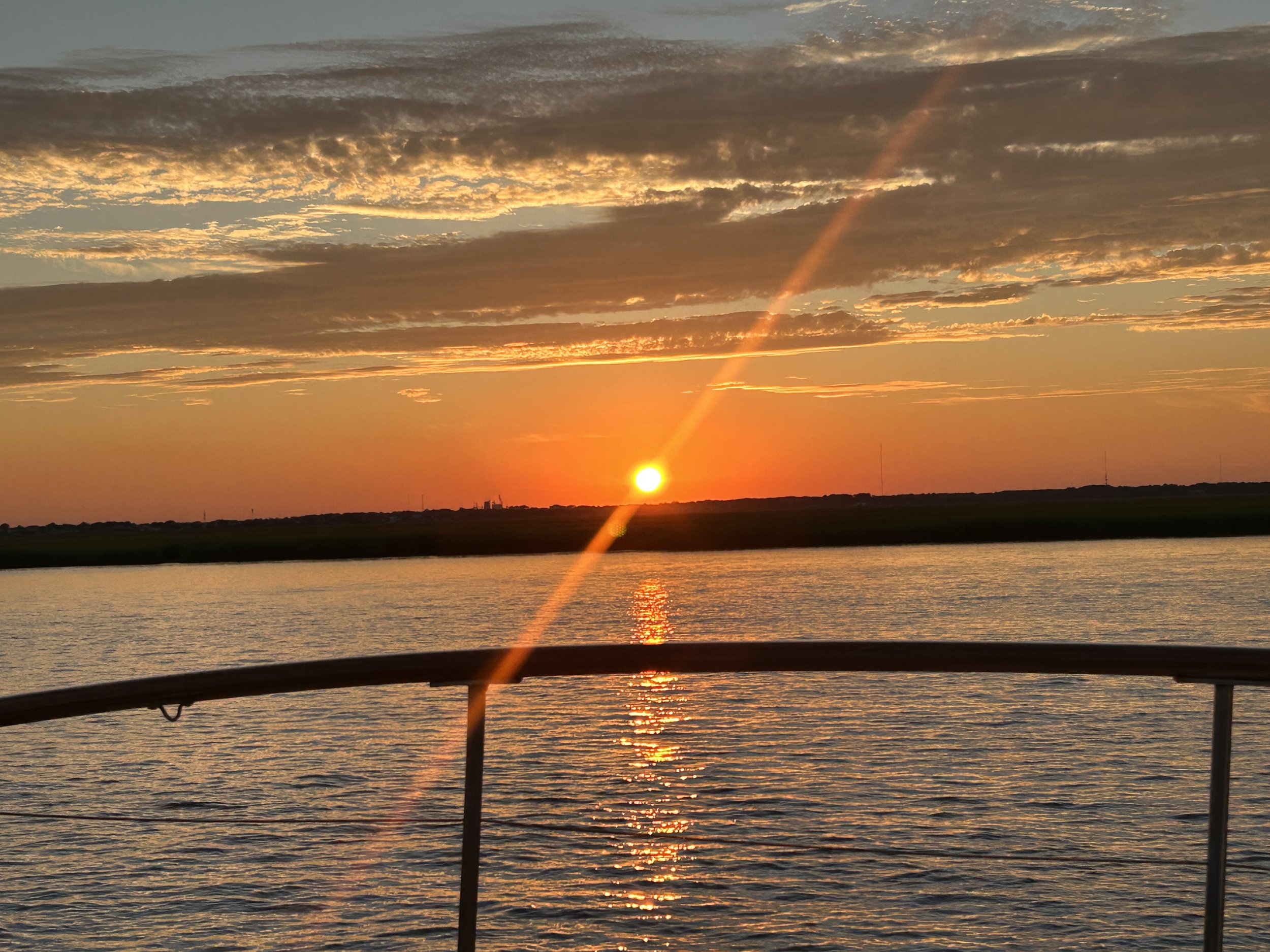 Sunset over a body of water with clouds in the sky and a faint rainbow, viewed from a boat with a metal railing in the foreground.