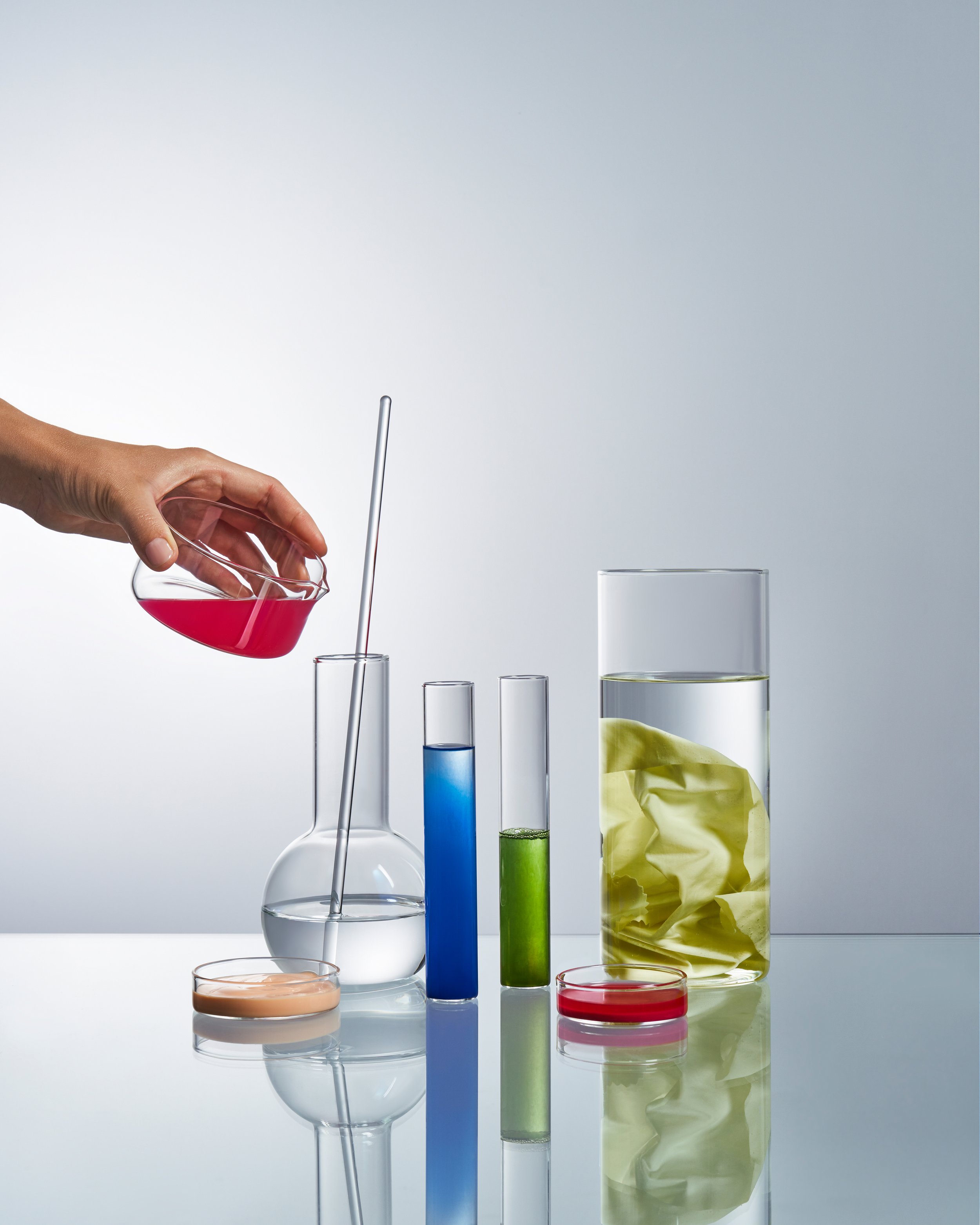 A hand pours a pink liquid from a beaker into a glass container on a white surface, with various test tubes and beakers filled with colorful liquids and a crumpled yellow paper in the background.
