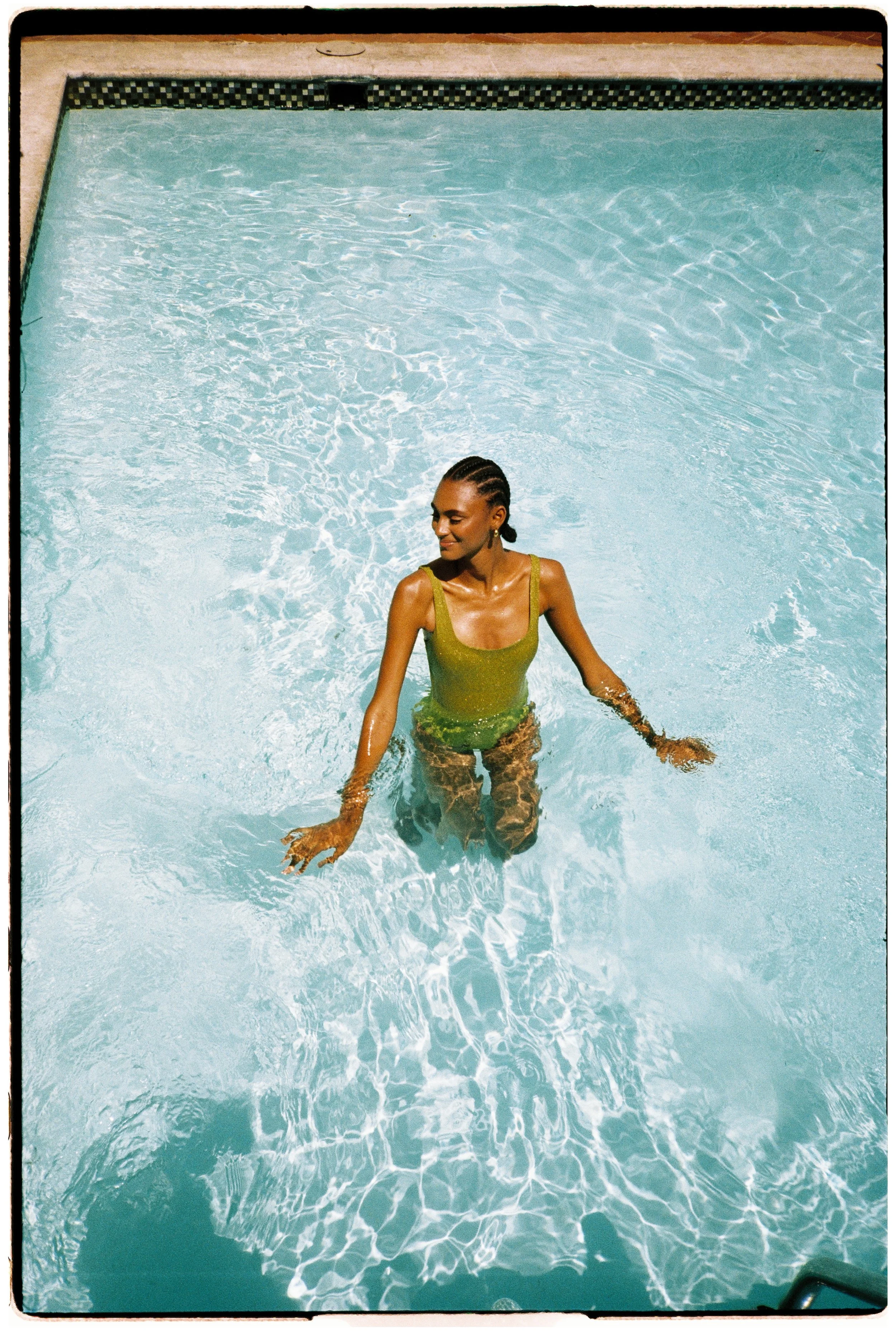 A woman with braided hair and gold earrings wearing a yellow swimsuit, standing in a swimming pool with clear blue water, smiling with her eyes closed.