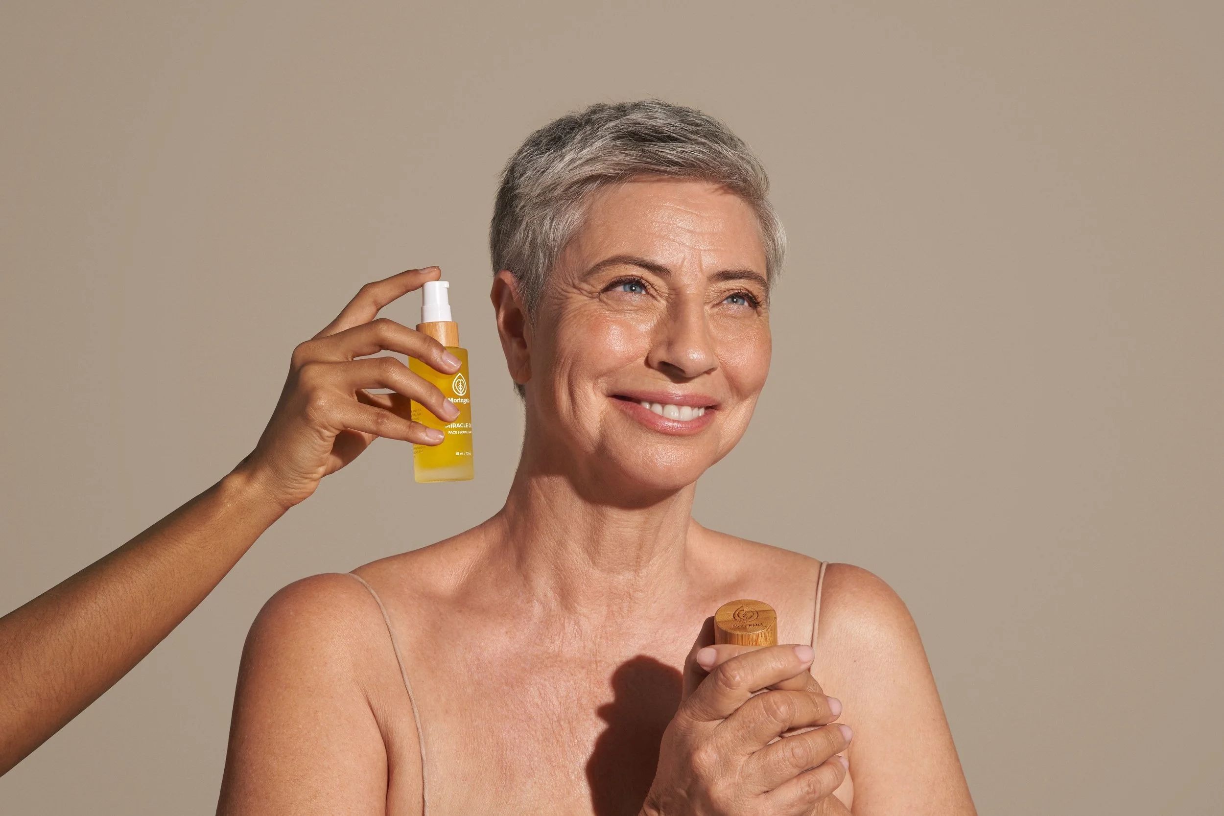 An older woman with short gray hair smiling while holding a wooden container. A hand is spraying her face with a yellow bottle labeled 'Moringa Miracle Oil'.