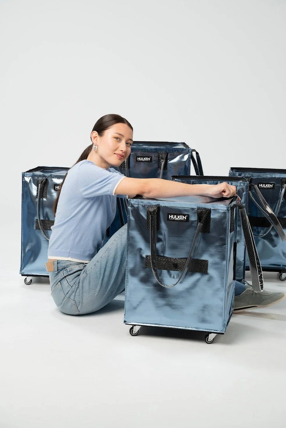 A woman sitting on the floor with her knees up, surrounded by large insulated delivery bags labeled "HULKEN." She is wearing a light blue top and jeans, and is reaching into one of the bags.