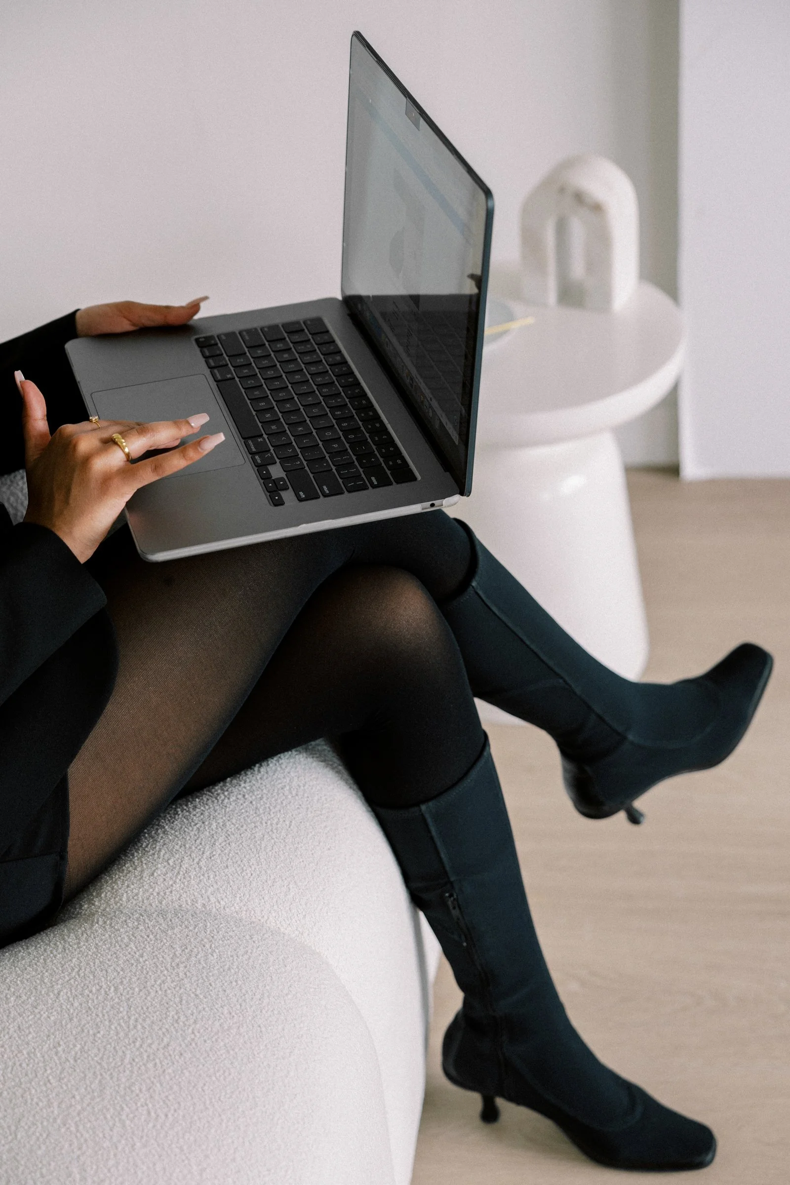 A woman sitting on a white couch using a silver laptop, wearing black boots and sheer black tights.