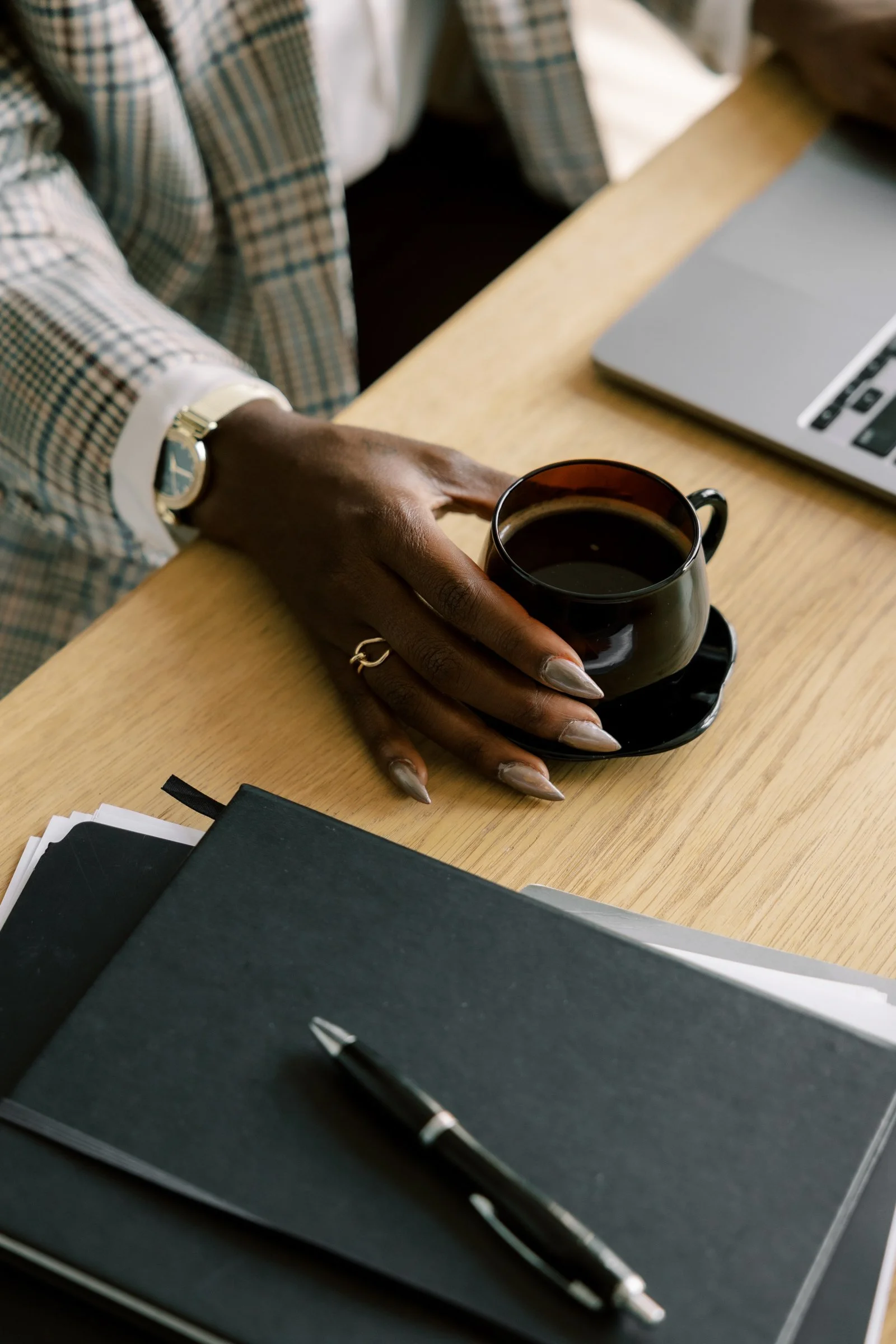 A person wearing a checkered blazer and a wristwatch is sitting at a desk, holding a black coffee cup on a black saucer. The desk has a laptop, some black notebooks, and a pen.