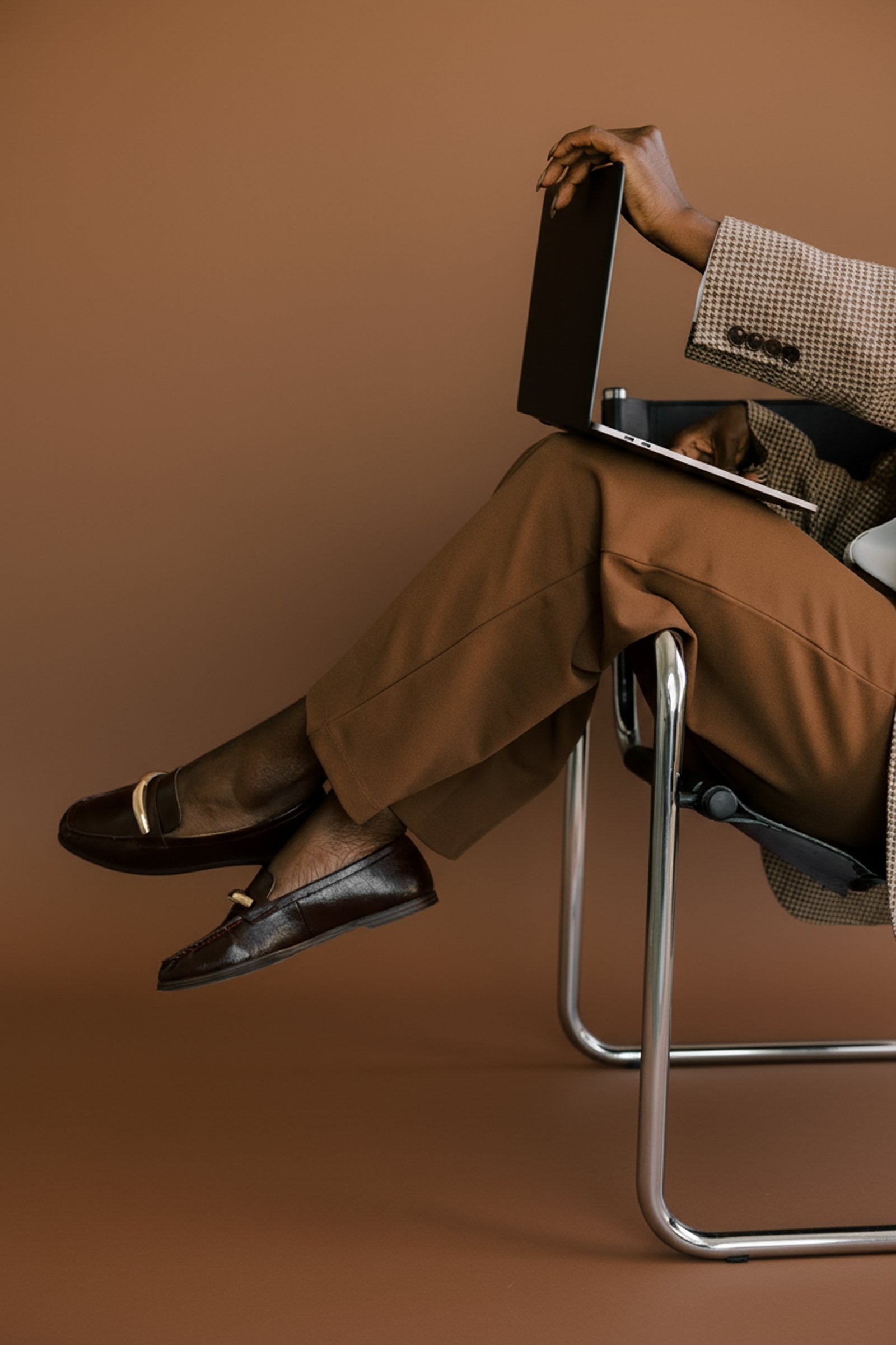 A person in brown trousers and dark brown shoes sitting on a modern metal chair with a brown background. The person is wearing a brown checkered blazer and is holding a closed laptop with the right hand.