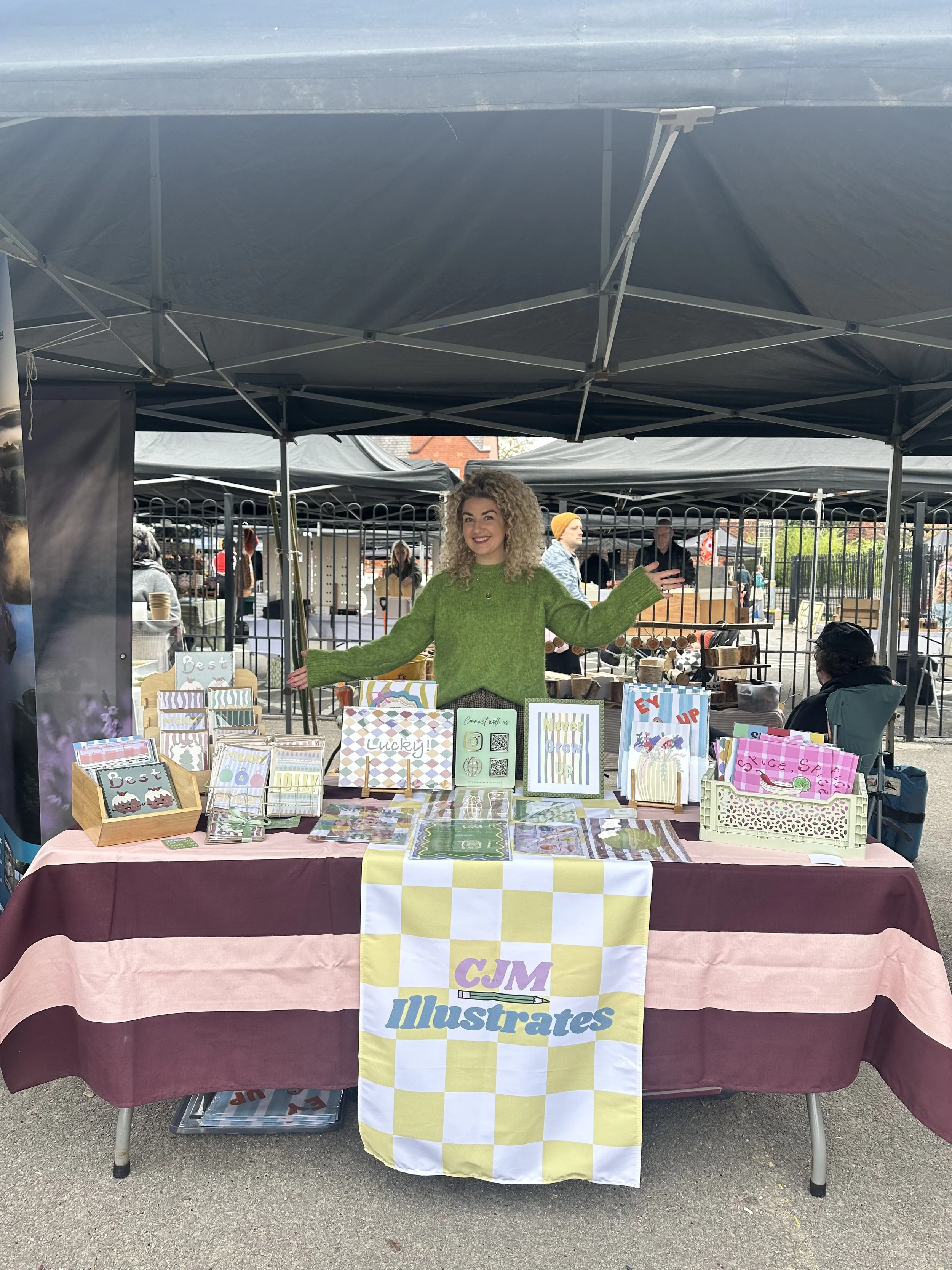A woman with curly blonde hair wearing a green sweater is standing behind a table at an outdoor market booth. The table displays colorful greeting cards and stationery items with various designs, and a banner that reads 'CJ M Illustrates.' The booth is set up under a black canopy, and there are other market booths and people visible in the background.