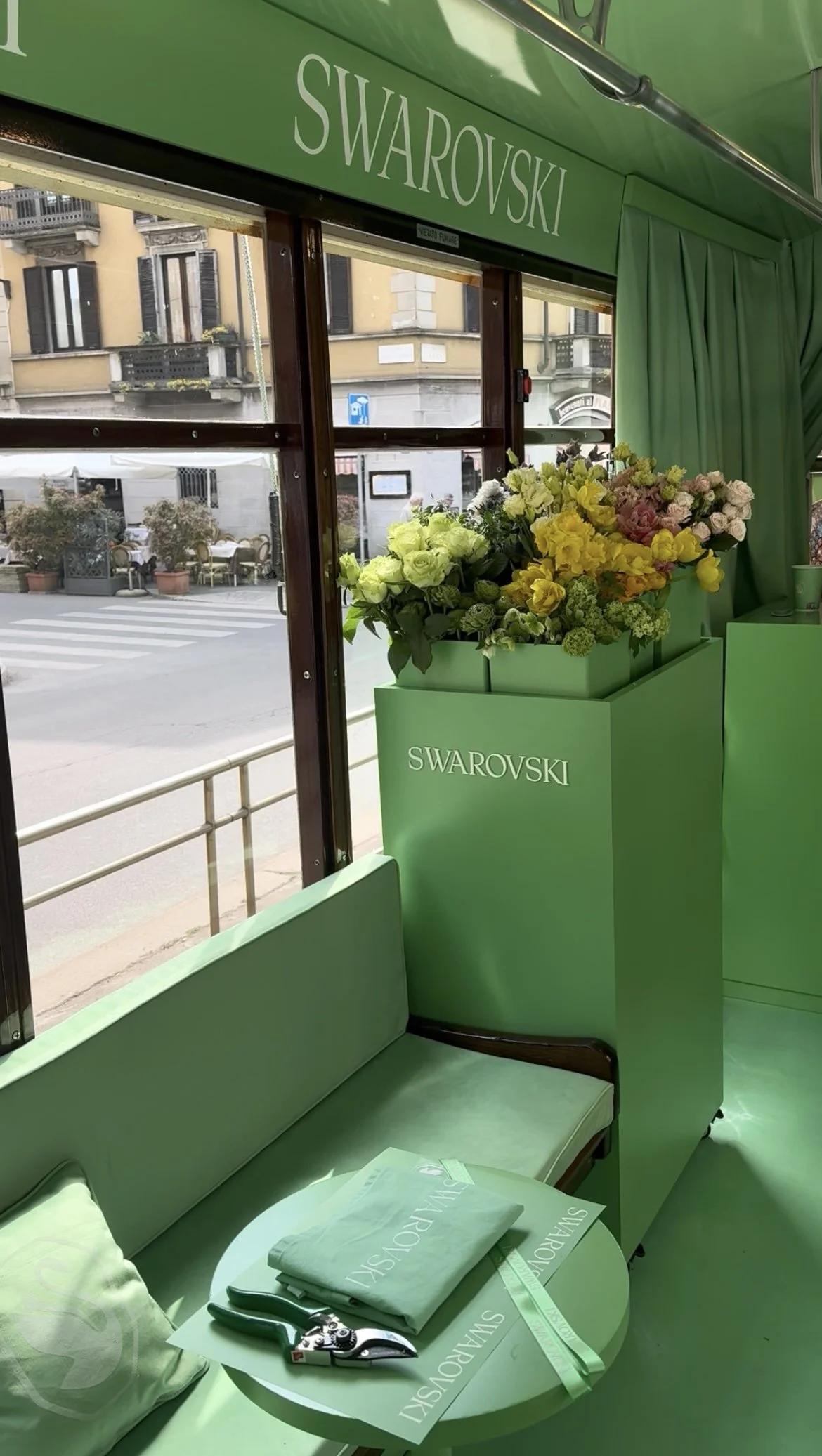 Interno di un vagone con decorazione verde, una pianta di fiori vari, sedile, tavolino con riviste, forbici e un lanyard, vista su strada con edifici e panchine fuori