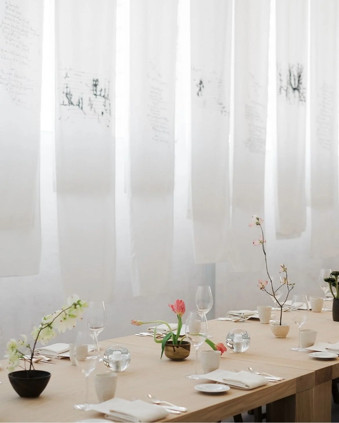 Tavola apparecchiata con bicchieri, piatti e tovaglioli, decorata con fiori di tulipano e orchidea in vasi, davanti a tende sintetiche con disegni tracciati.