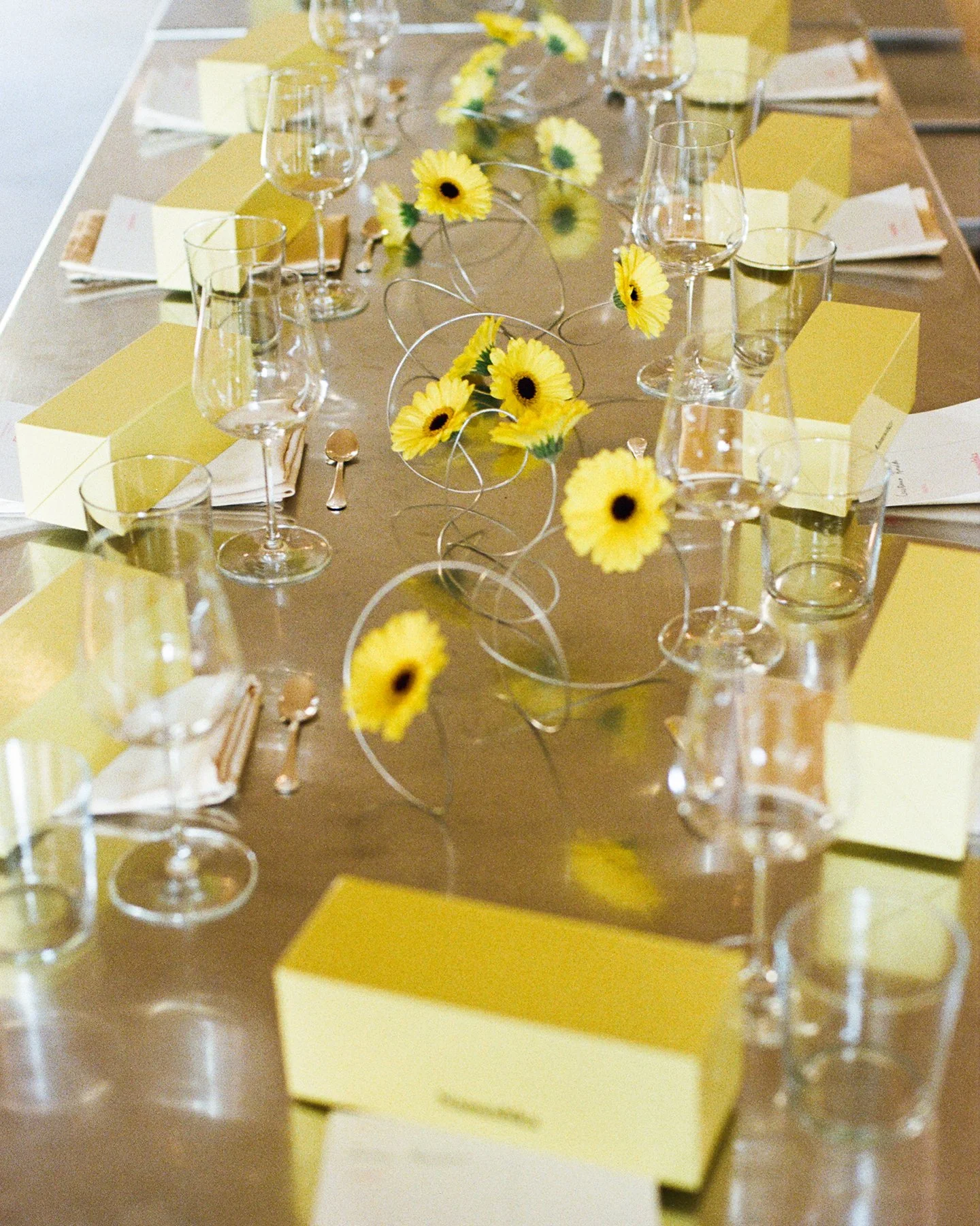 Tavola apparecchiata con bicchieri di vino, fiori gialli e pezzi di carta color giallo, pronto per un evento.