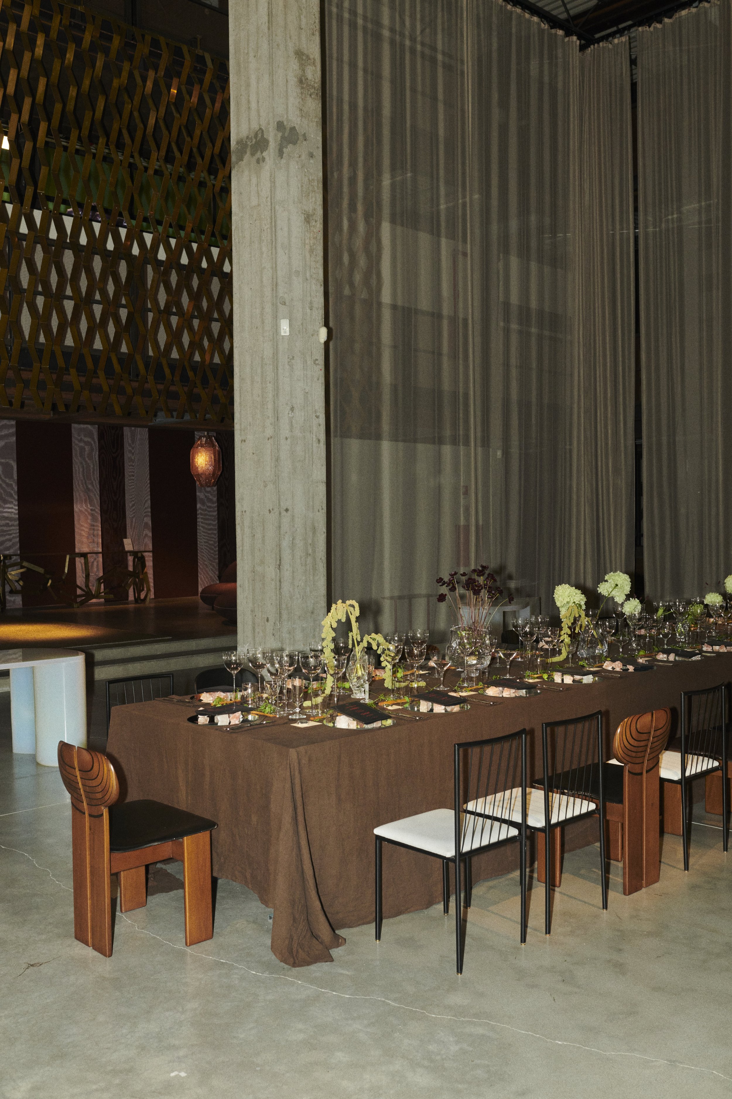 Tavolo apparecchiato con tovaglia marrone, fiori e cristalli in un ristorante elegante, sfondo con drappi di tessuto e decorazioni di design moderni.