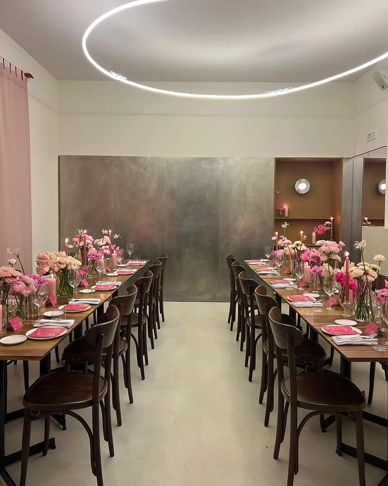 Sala da pranzo elegante con due lunghe tavole rettangolari, decorate con fiore all'occhiello di peonie rosa e altri fiori rosa, sono apparecchiate con piatti, bicchieri e posate. Le sedie sono in legno scuro e le luci sospese sul soffitto creano un'atmosfera moderna.