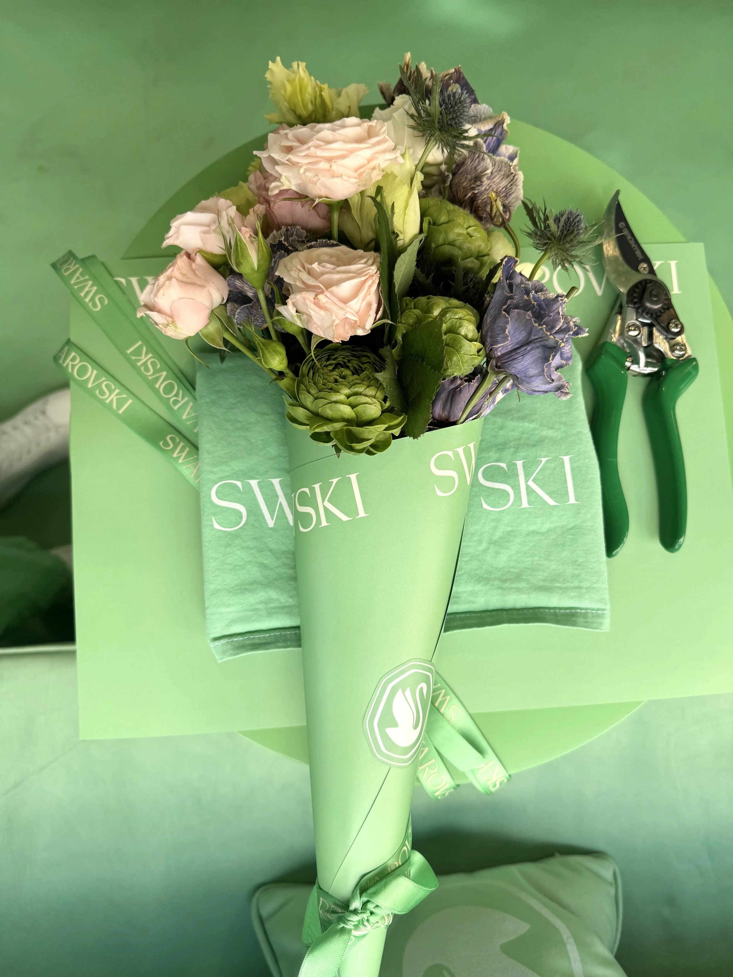Un mazzo di fiori misti, tra cui rose rosa pallido e fiori viola, avvolti in una carta verde con il logo 'Suzuki' in bianco, viene mostrato. Lo sfondo è di un pannello verde.