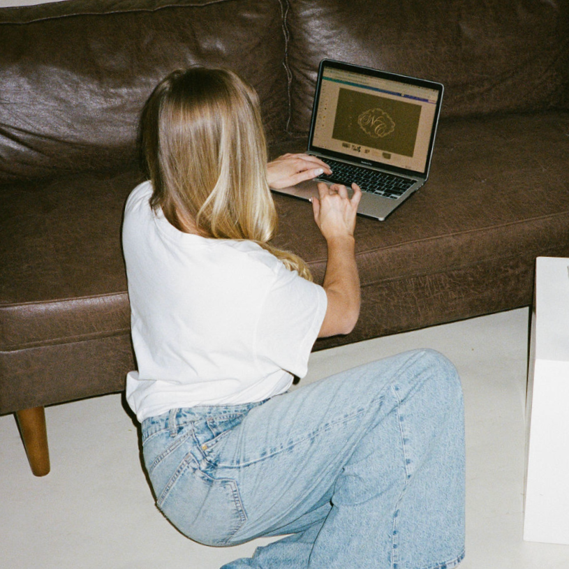 A woman with blonde hair wearing a white T-shirt and light blue jeans sitting on the floor next to a brown sofa, using a laptop.