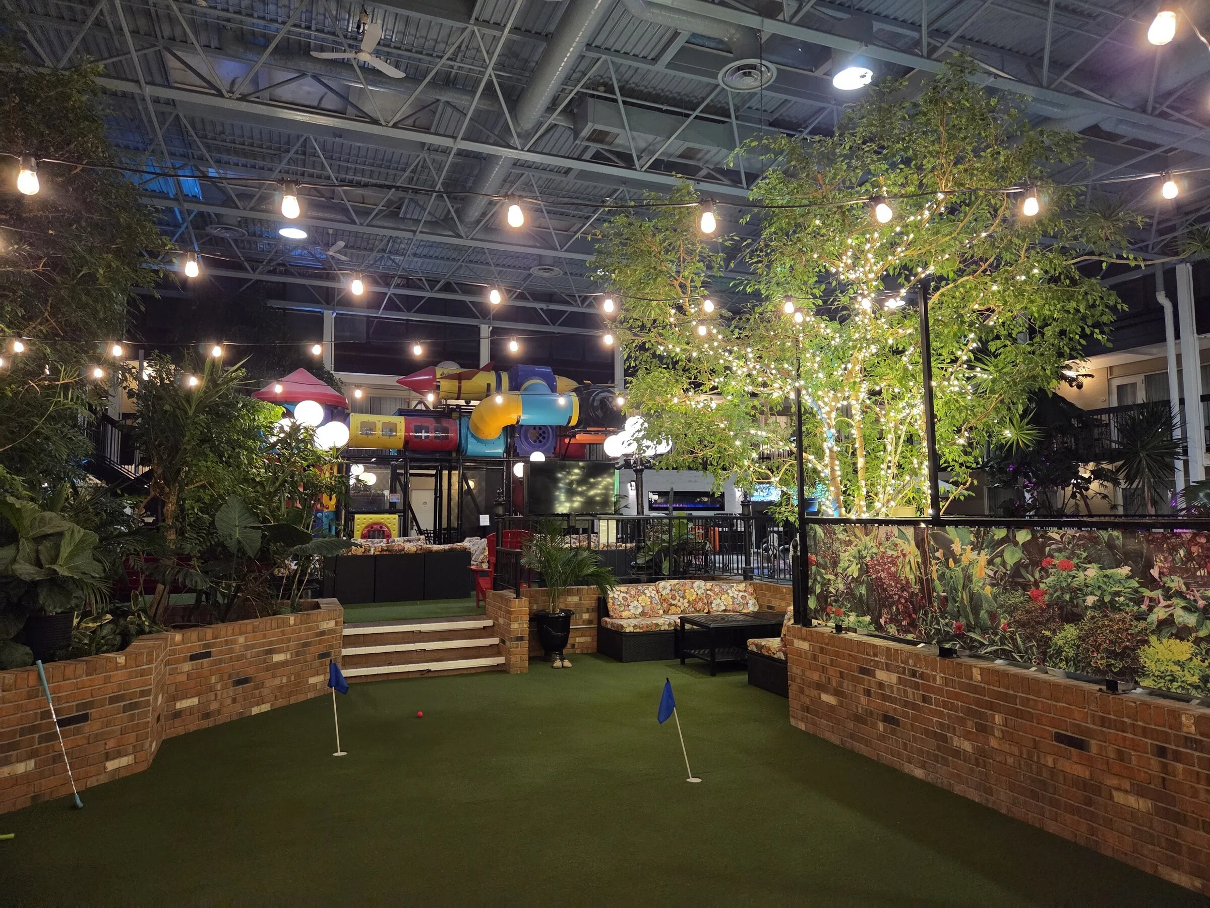 Commercial indoor atrium with parallel bistro light runs over putting green, lounge seating, and playground zone.