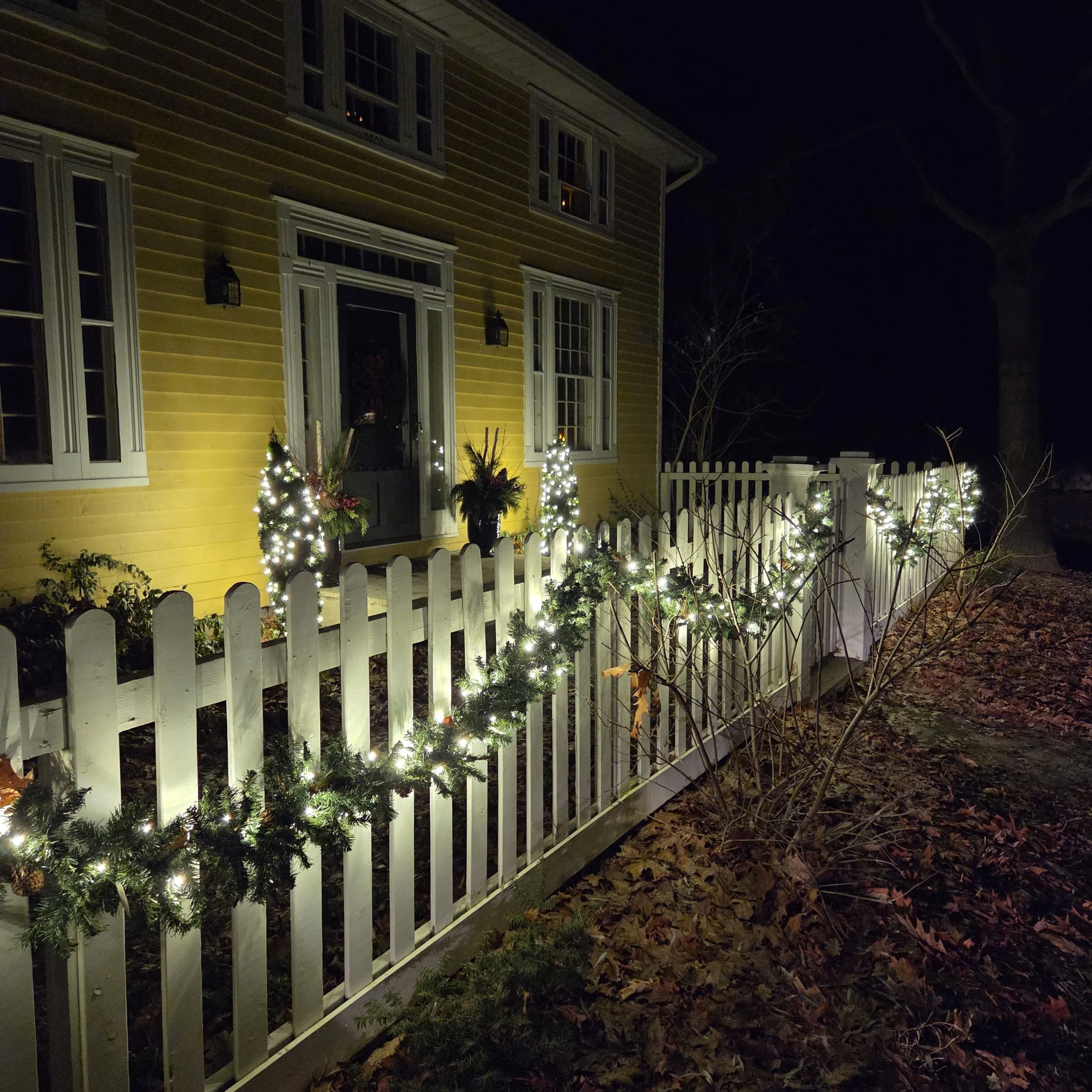 Niagara-on-the-Lake home at night with red and white C9 roofline lights, warm-white bush wraps, and a lit wooden fence after light snowfall.