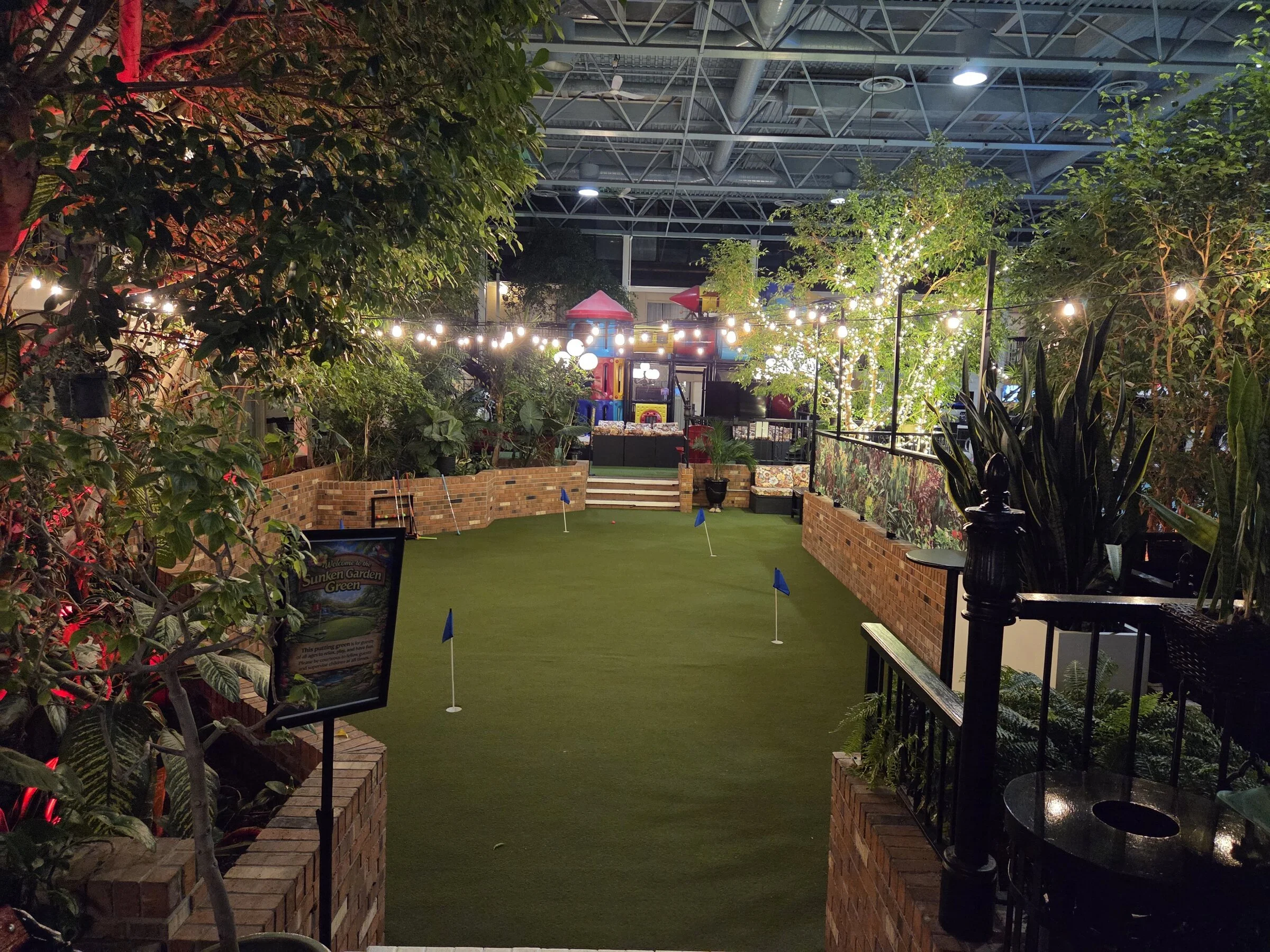 Warm Edison-style permanent bistro lights strung over an indoor putting green atrium with brick planters and lush foliage in Niagara Falls.