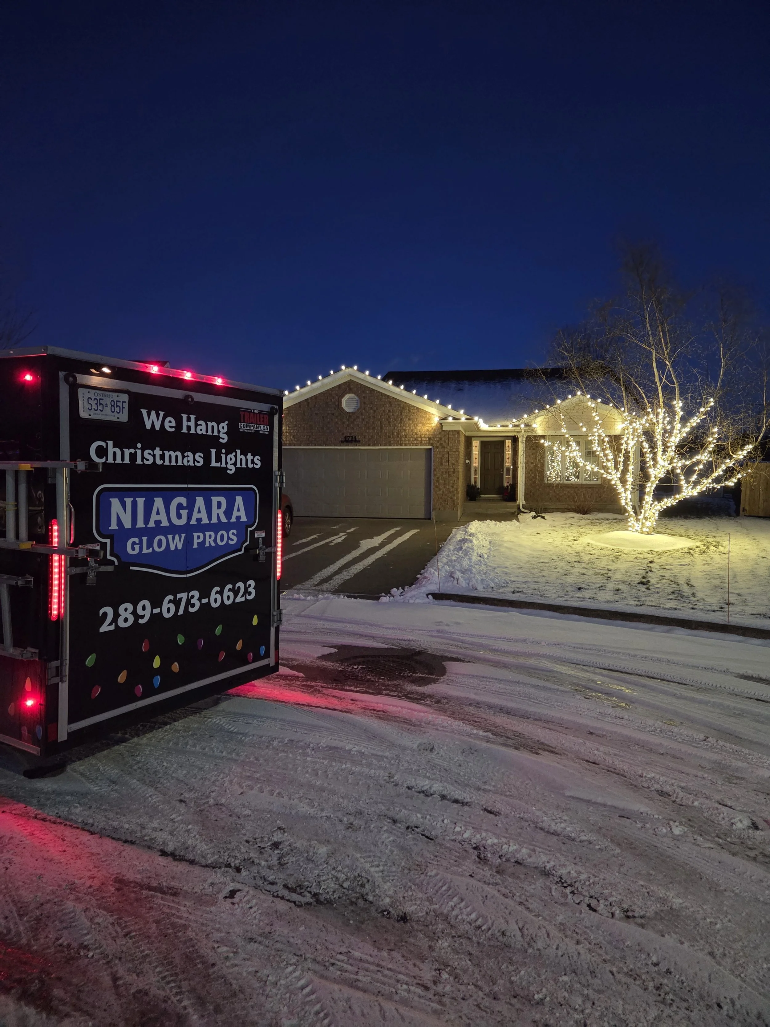 Niagara Glow Pros branded trailer at a Niagara Falls residential install with white roofline lights and a warm-white wrapped tree.