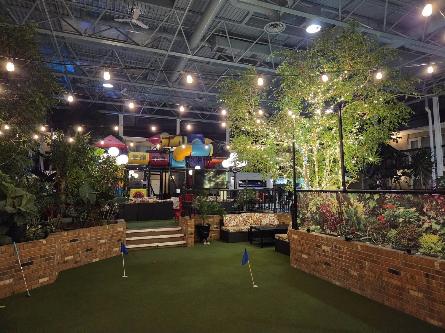 Commercial indoor atrium with parallel bistro light runs over putting green, lounge seating, and playground zone.