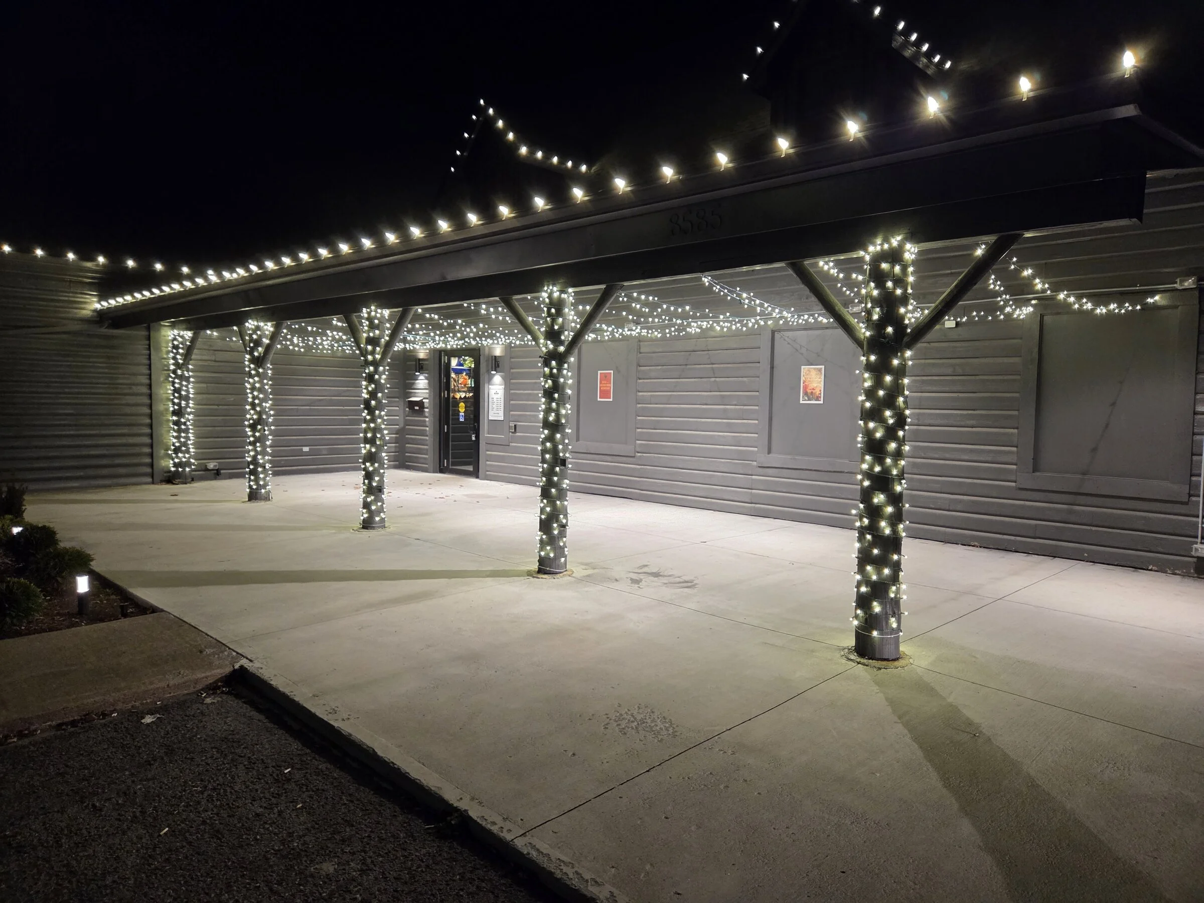 Commercial porch pillars and awning outlined in warm-white Christmas lights at night.