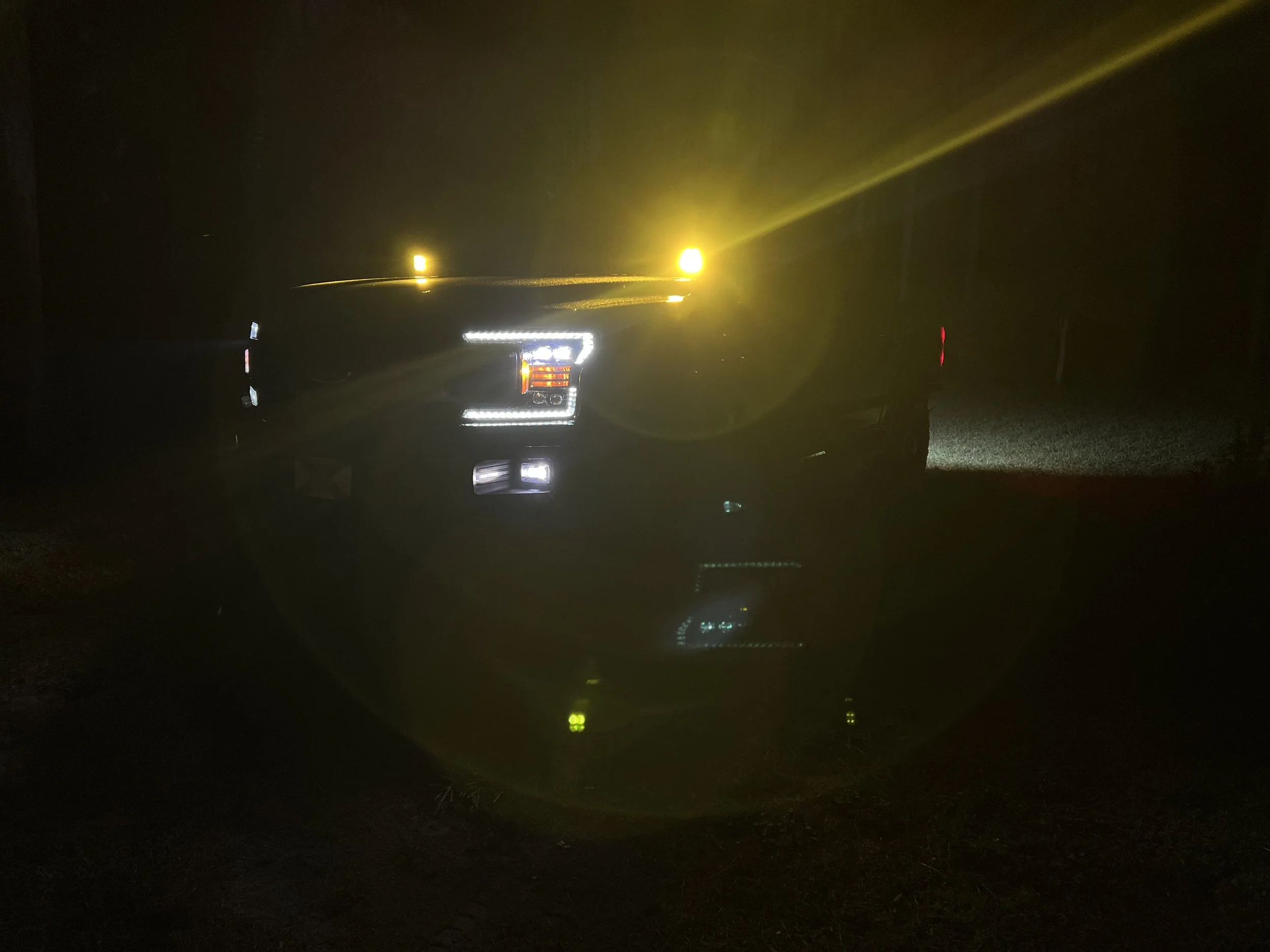 A dark night scene showing a vehicle with illuminated headlights and yellow warning lights, with the light reflecting off a mirror surface.