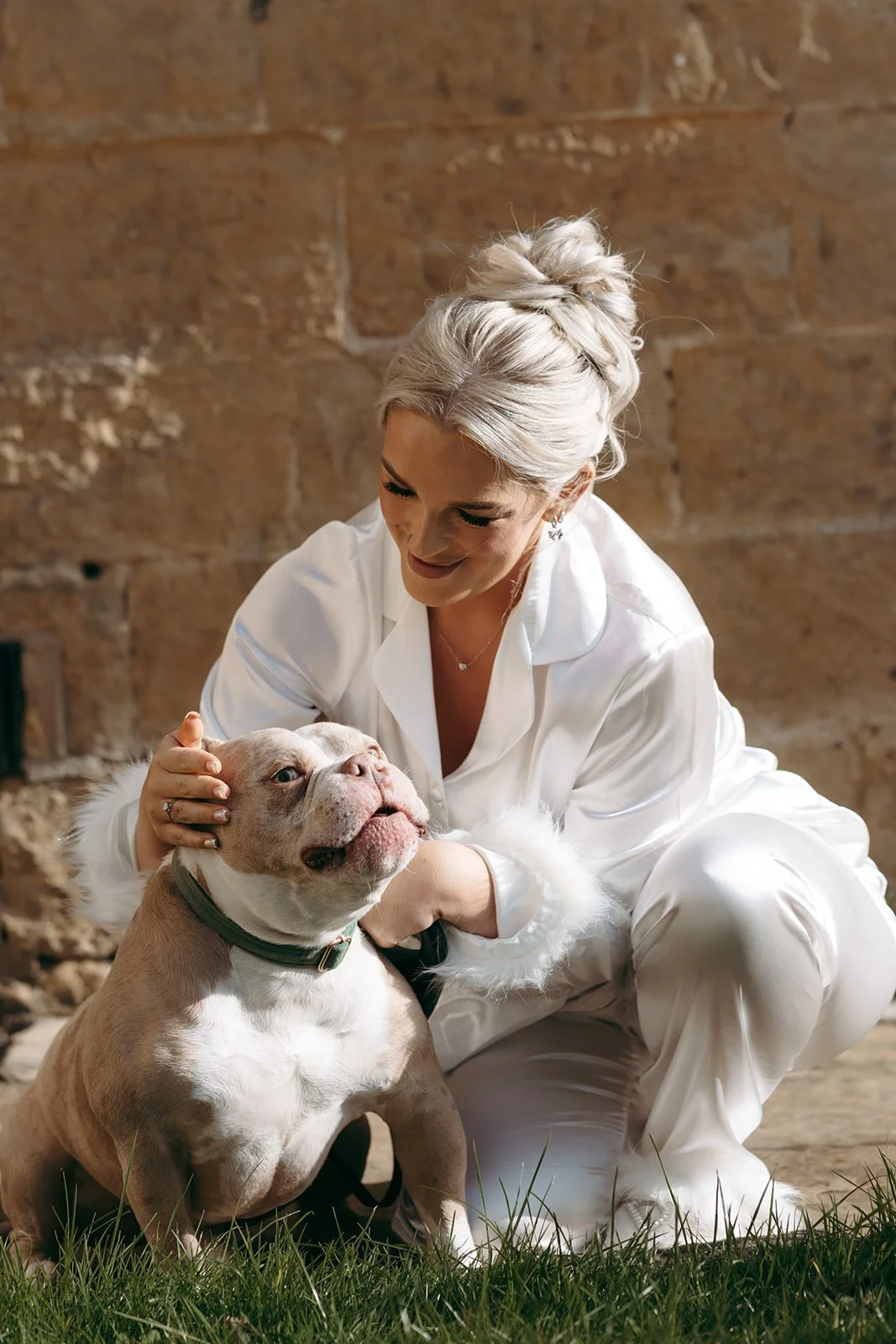 A woman with platinum blonde hair in an updo, wearing white satin pajamas, crouches on grass and gently holds a large American Bully dog, smiling down at it. The dog is light brown and white with a black collar, sitting on the grass with a relaxed ex