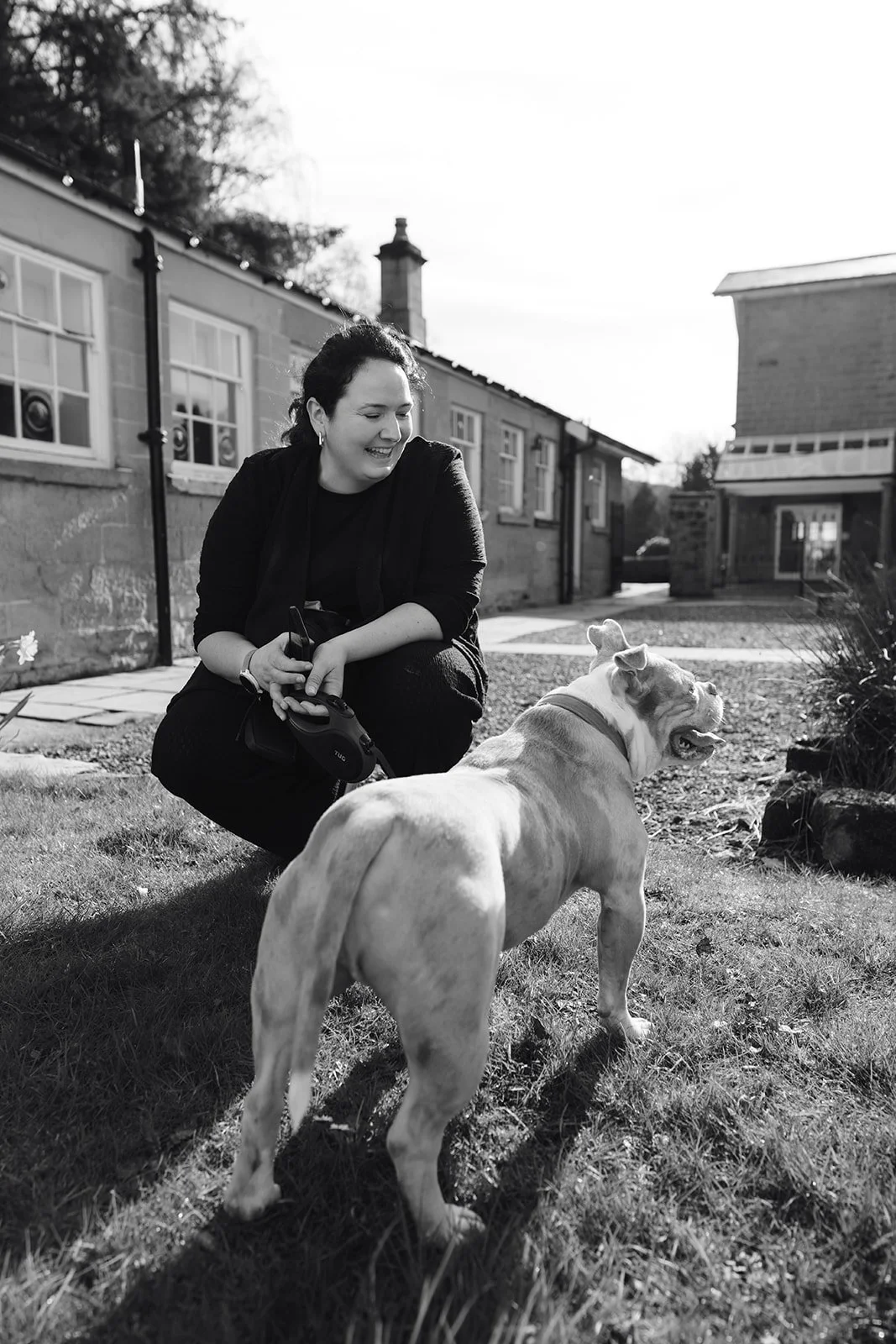 A woman with dark hair crouching and smiling at a dog in a yard on a sunny day, with a house in the background.