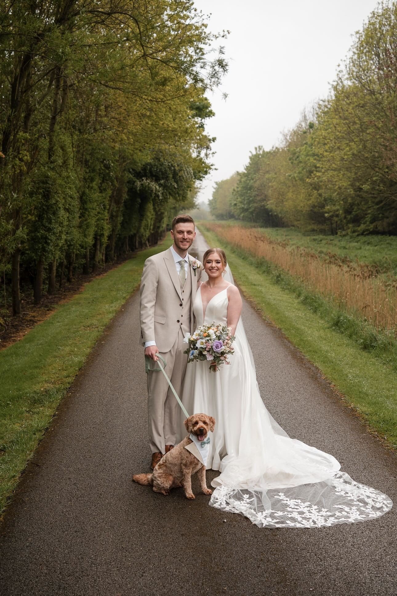 Bride and groom smiling with beautiful trees behind them with their dog standing at their feet with a bow tie on