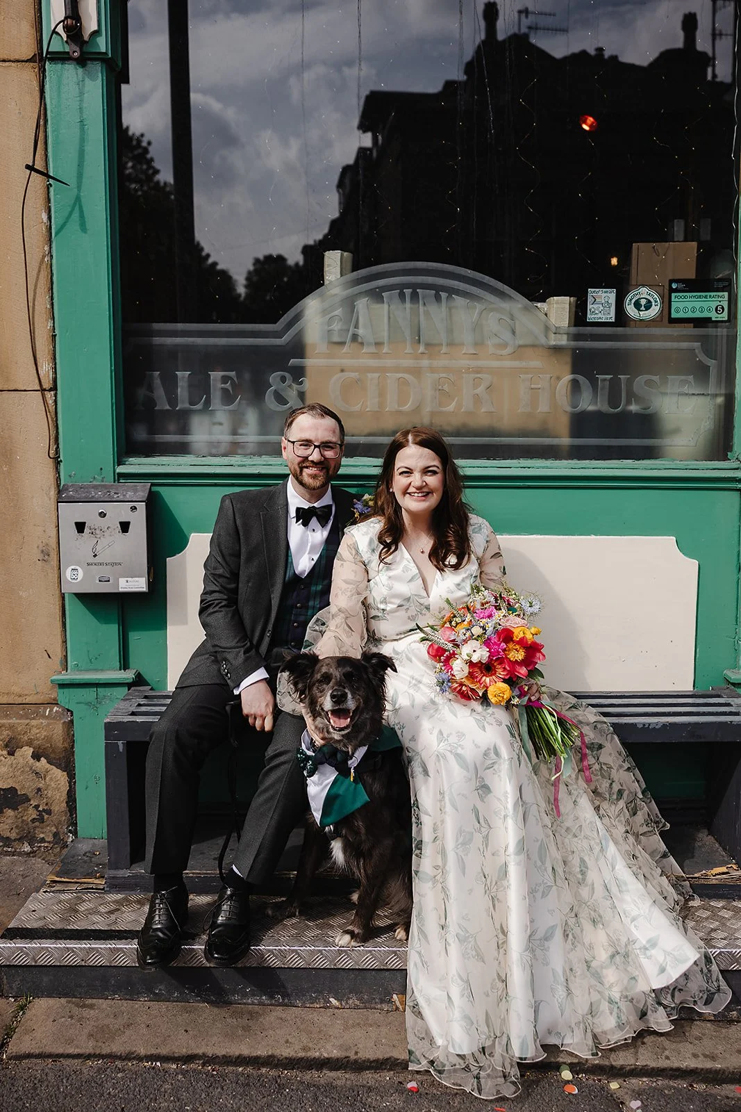 A bride and groom sit on a bench outside a pub with their dog sat between them, wearing a green tux and bow tie.