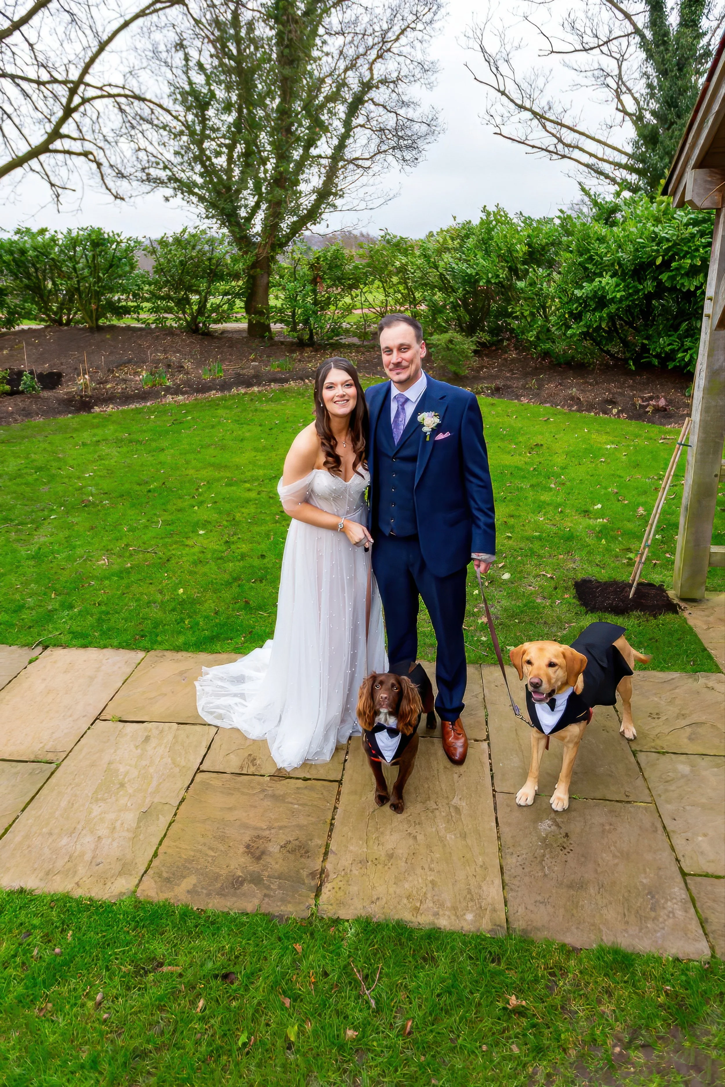 A bride and groom standing outdoors on a stone patio, dressed in wedding attire, accompanied by two dogs in tuxedos. The bride wears a white strapless wedding dress, and the groom wears a navy blue suit with a purple tie and boutonniere. The background features a garden with green grass, shrubs, and trees. The sky is cloudy.