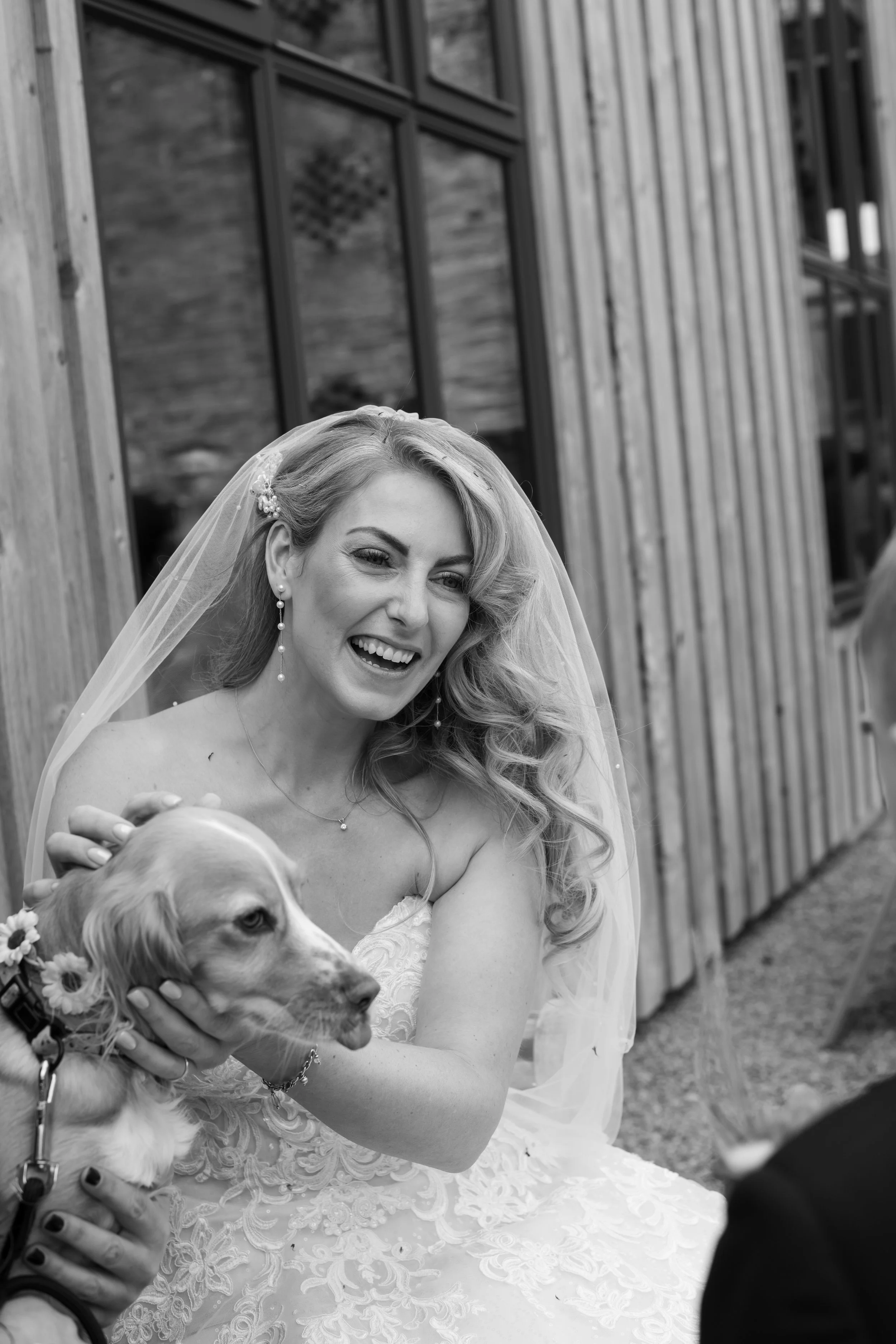 Black and white photo of a woman in a wedding dress and veil, smiling and petting a dog with flowers around its neck, outside a building with wooden siding and large windows.