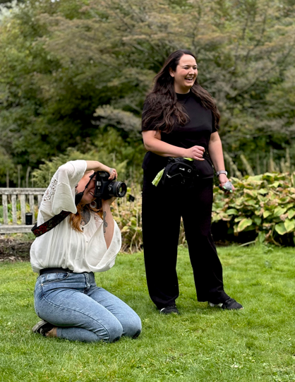 A woman with long hair, wearing a black shirt and black pants, smiling and standing outdoors on a grassy lawn. Another woman, kneeling and wearing a white shirt and blue jeans, is taking a photo or video of her with a camera.