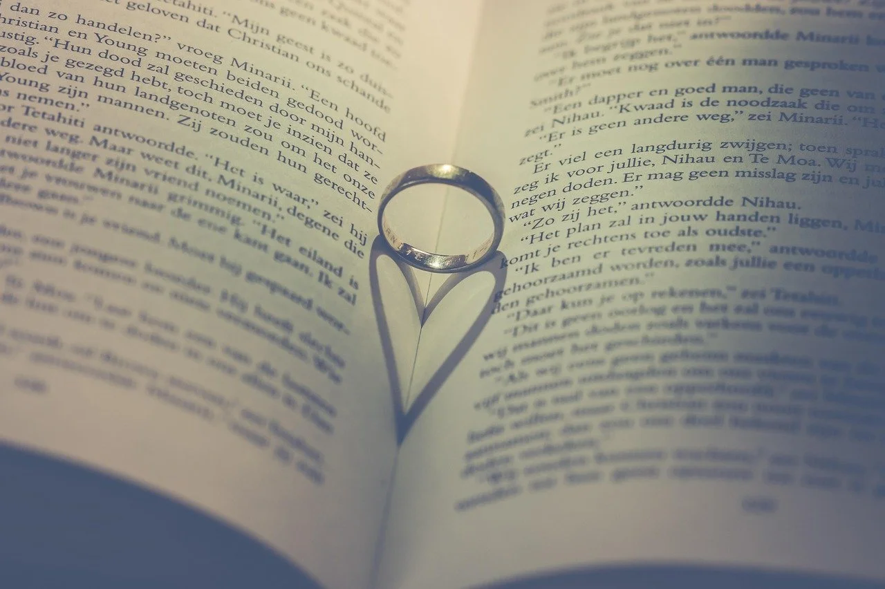 A silver ring placed on an open book creates a heart-shaped shadow of the ring's reflection on the page.