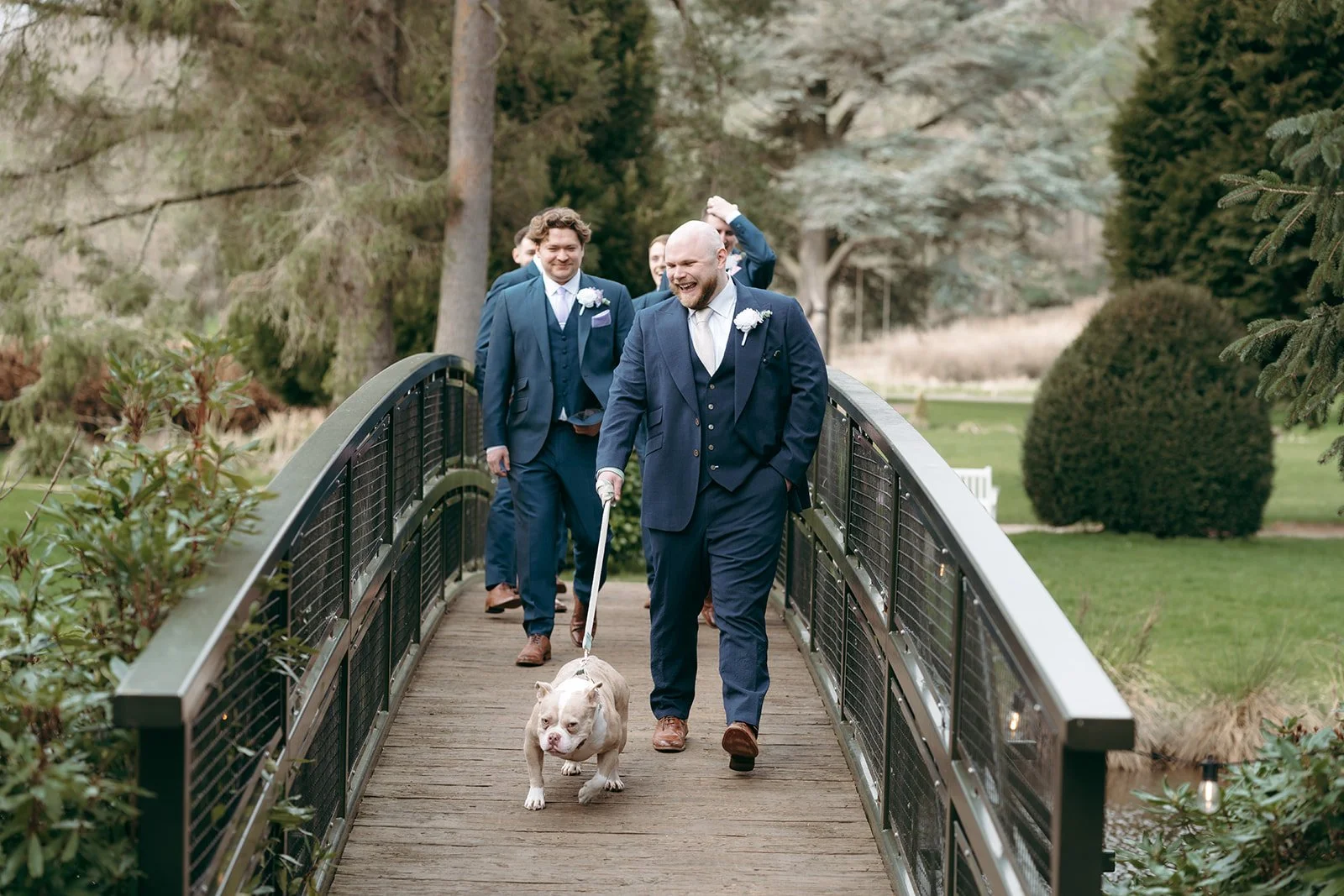 A man in a navy suit leading a dog across a small bridge, followed by several men in suits, in a park-like setting with trees and bushes.